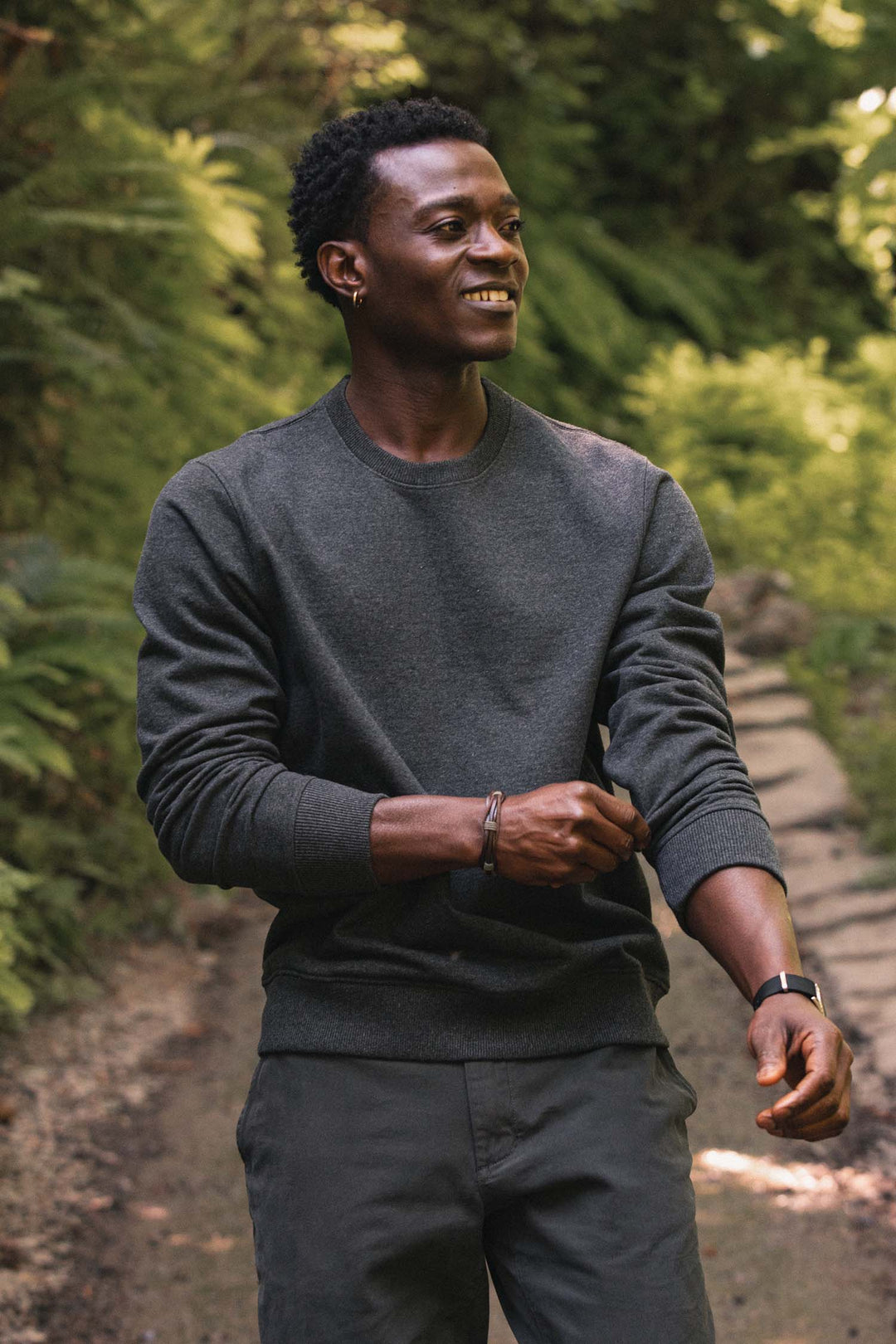 A person wearing the Bridge & Burn Hayden Crew Sweatshirt in Graphite and matching pants stands on a nature trail, smiling as they roll up their sleeve, surrounded by lush green trees and plants.