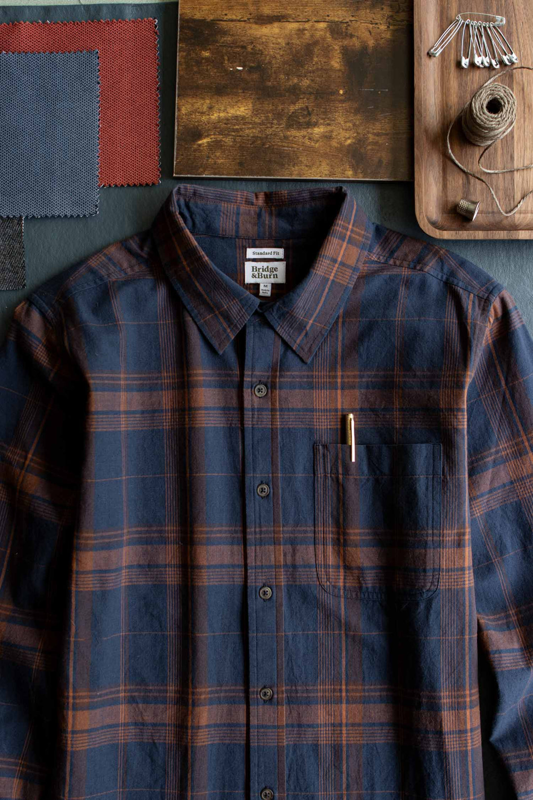 The Bridge & Burn Larson Shirt in Cinder Plaid—a classic fit, navy and brown brushed flannel button-up—features a pen in its chest pocket and is styled alongside fabric swatches, a wooden board, and a tray containing safety pins and twine.