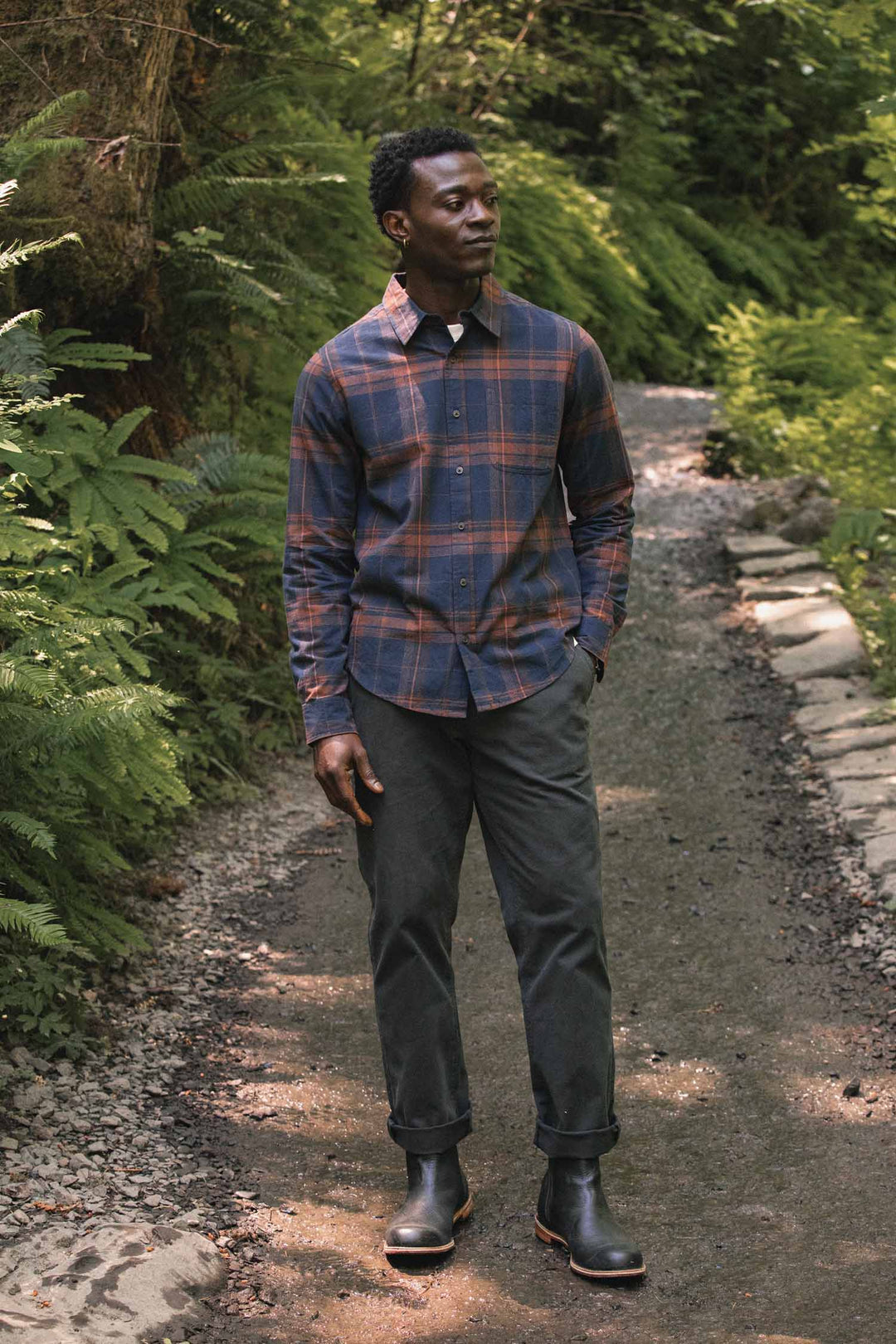 A man stands on a forest path, wearing the Bridge & Burn Larson Shirt in Cinder Plaid with dark pants and black boots. Lush green foliage surrounds him, and a stone border lines the trail.