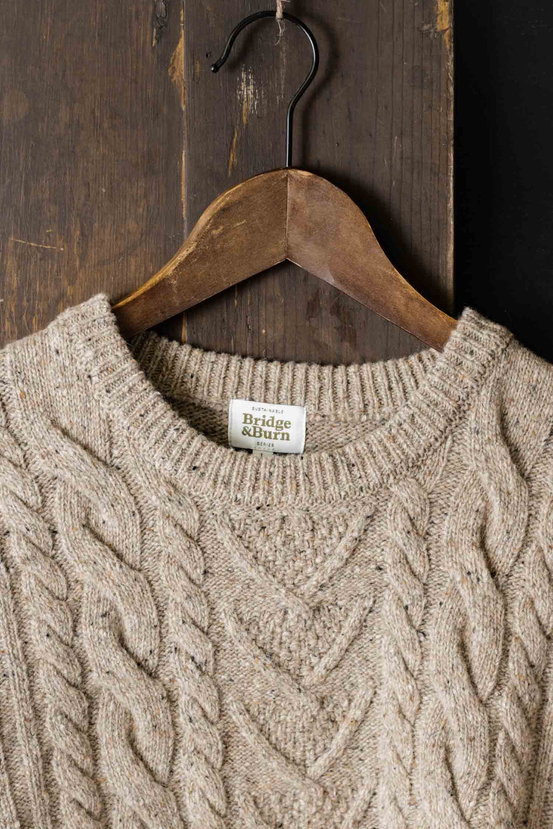 A flecked oatmeal Ludlow Sweater by Bridge & Burn, crafted from RWS-certified wool, hangs on a wooden hanger against a dark wood backdrop. The ribbed collar and ethical materials highlight quality craftsmanship.