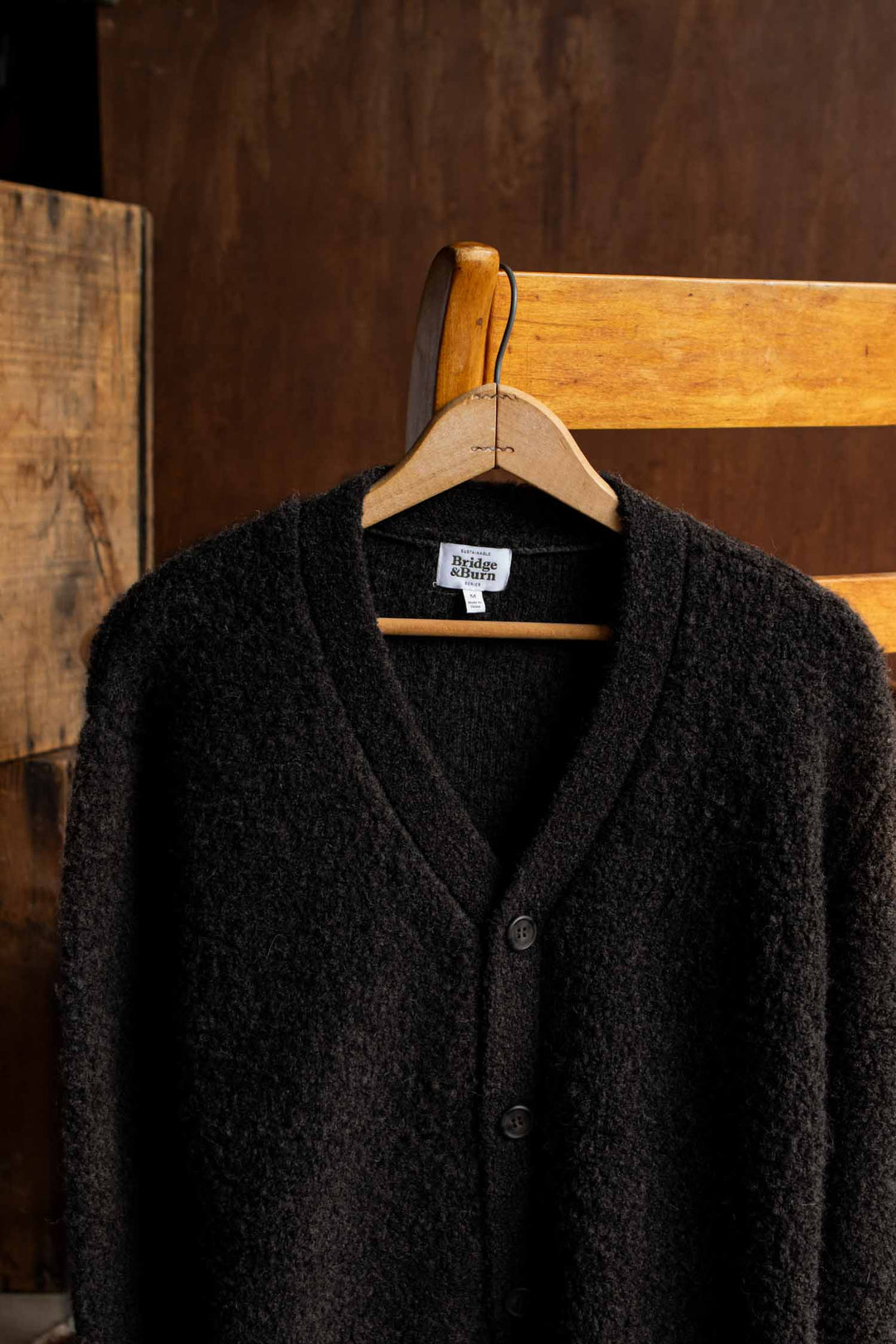 The Bridge & Burn Mateo Cardigan in Black, featuring a dark textured knit, V-neckline, button closure, and patch pockets, hangs on a wooden hanger draped over a wooden chair set against a brown background.