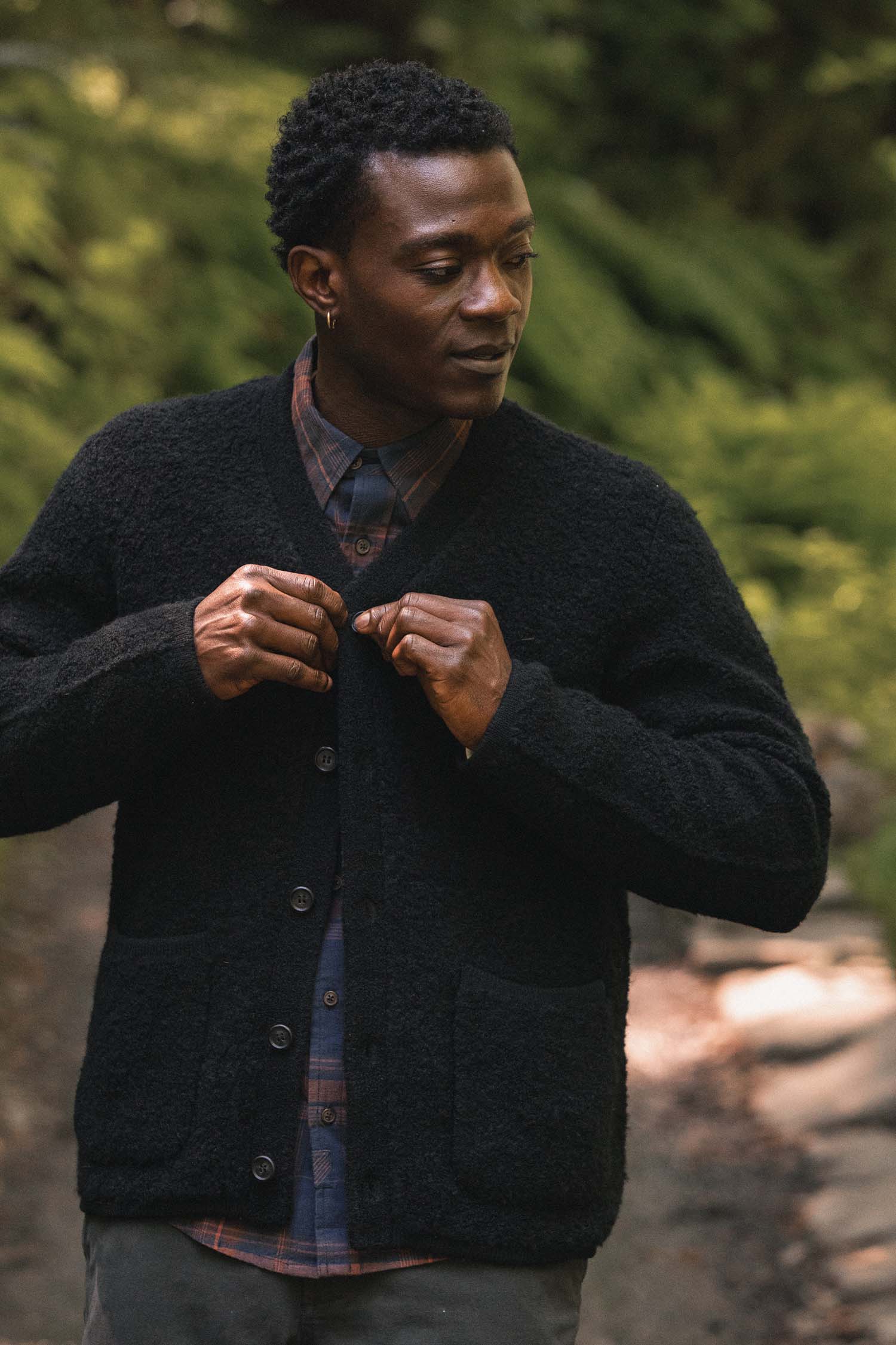A person stands outdoors on a path, looking to the side while buttoning their Bridge & Burn Mateo Cardigan in Black—a textured knit V-neck sweater with patch pockets—layered over a plaid shirt, with blurred greenery in the background.