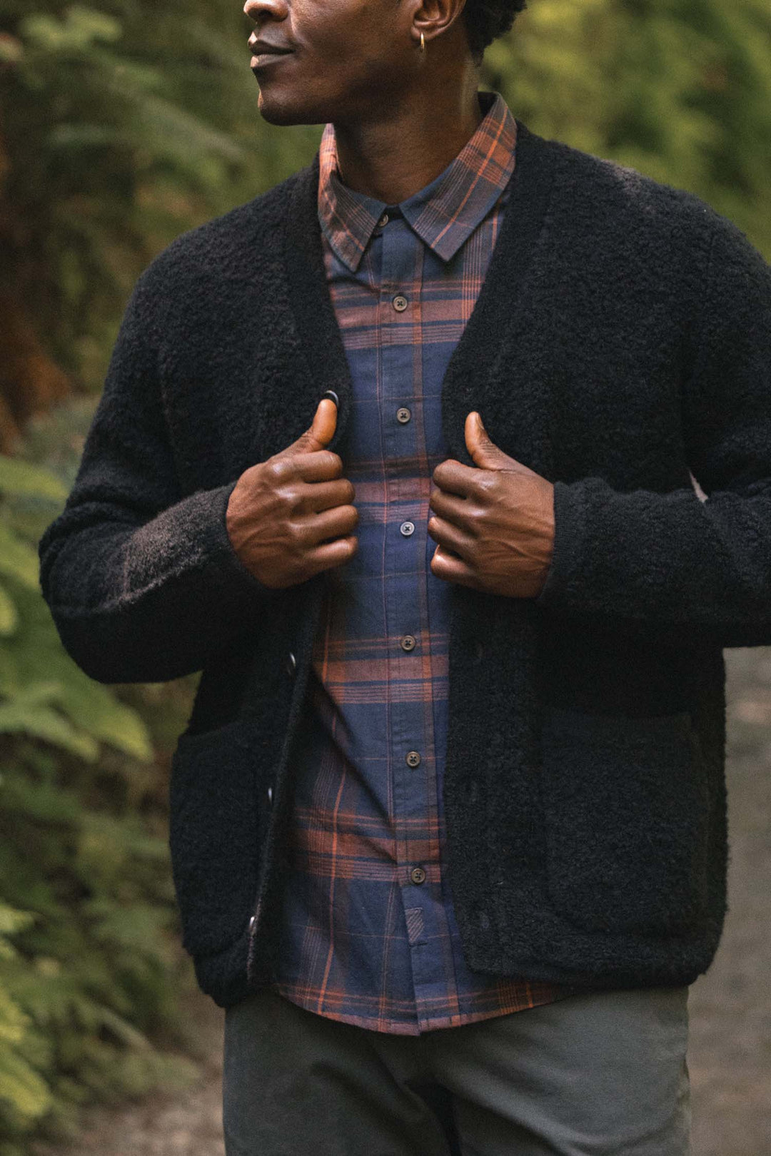 A person stands outdoors wearing the Bridge & Burn Mateo Cardigan in black—a dark, textured knit V-neck cardigan with patch pockets—layered over a blue and red plaid shirt and holding the cardigan edges with both hands. The background features greenery and a pathway.
