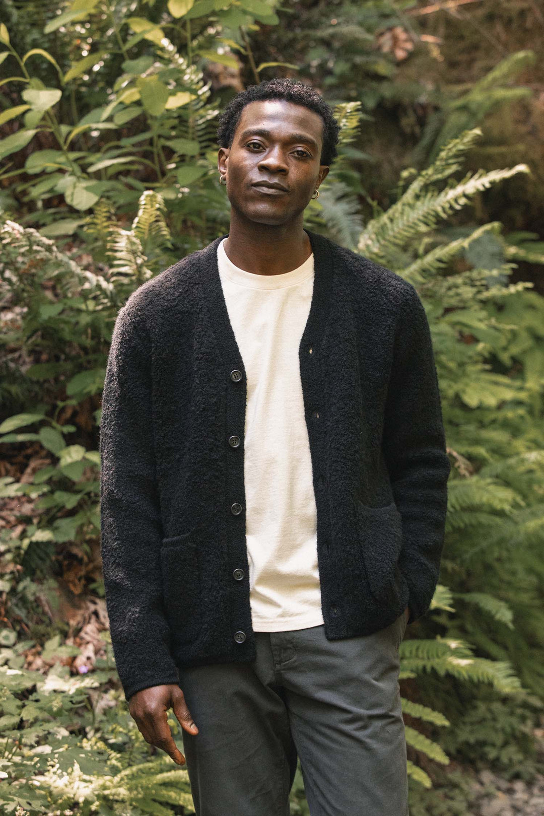 A man stands outdoors surrounded by green foliage, wearing a white shirt, gray pants, and the Bridge & Burn Mateo Cardigan in black—a dark textured knit V-neck sweater with patch pockets—looking calmly at the camera with one hand in his pocket.