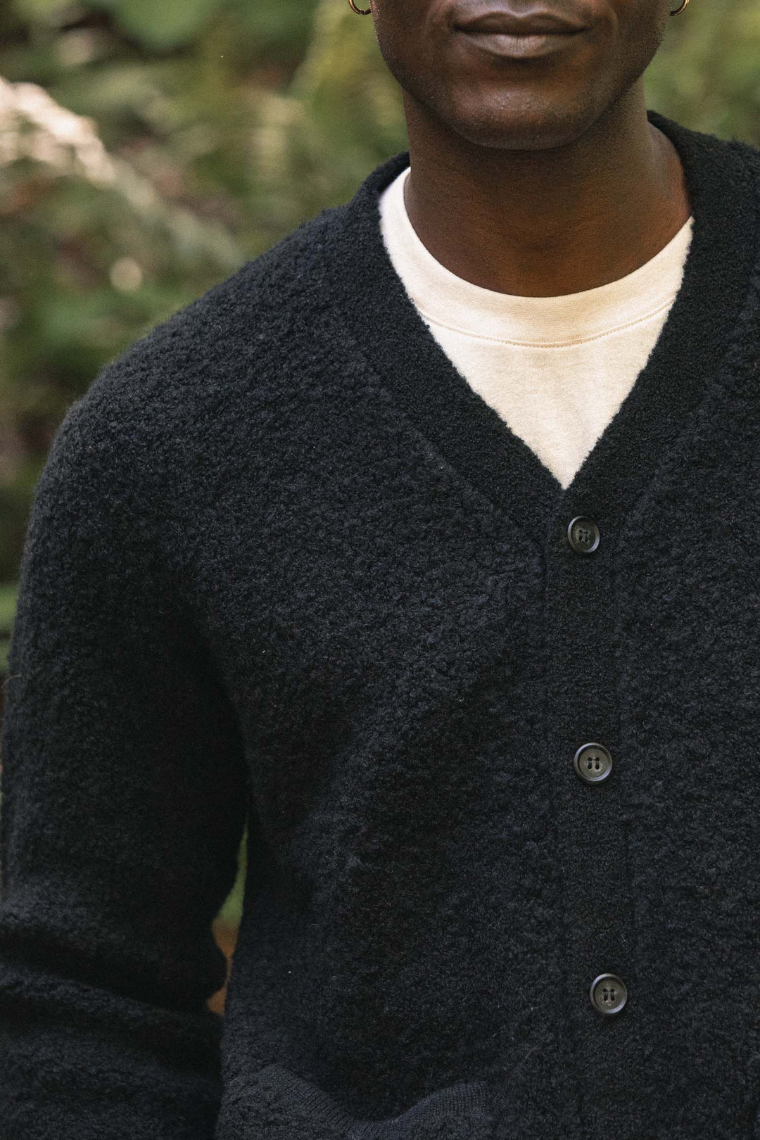 A person wearing the Bridge & Burn Mateo Cardigan in black—a textured knit V-neck style with patch pockets—layered over a white shirt stands outdoors, greenery softly blurred in the background. Only the lower half of their face and upper torso are visible.