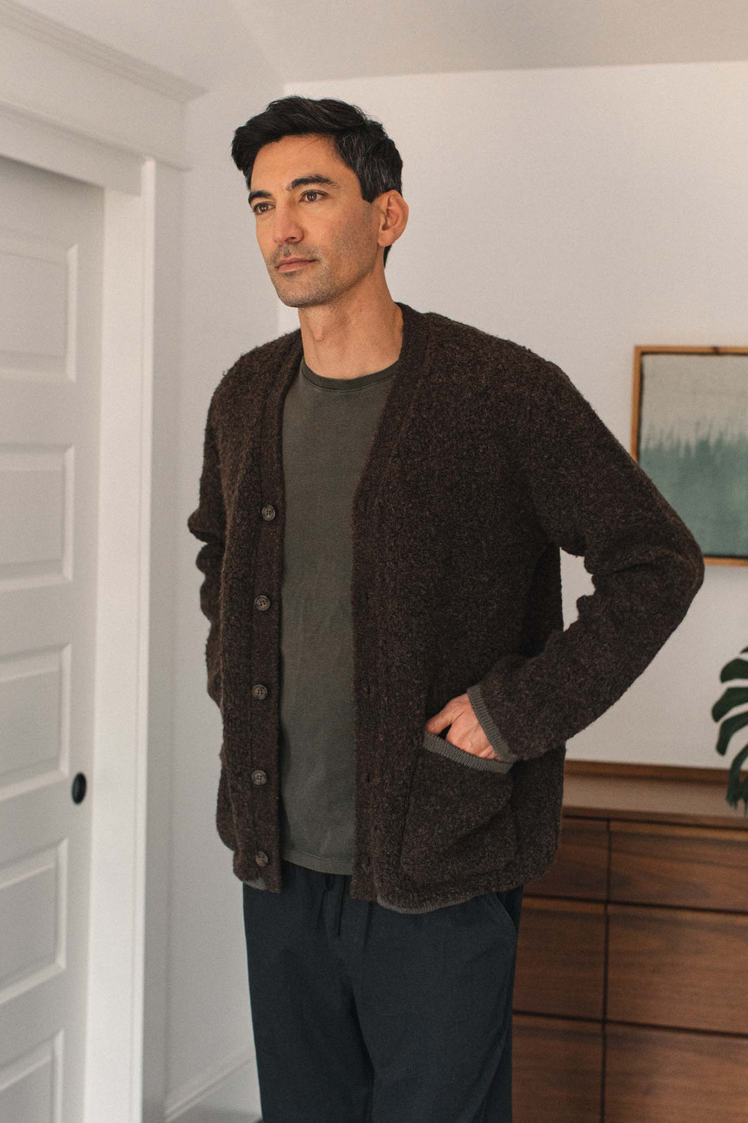 A man with short dark hair stands indoors, wearing the Bridge & Burn Mateo Cardigan in Dark Olive—a textured knit V-neck style with patch pockets—over a gray shirt and dark pants, his hands resting in his pockets. A dresser, a plant, and a framed picture can be seen in the background.