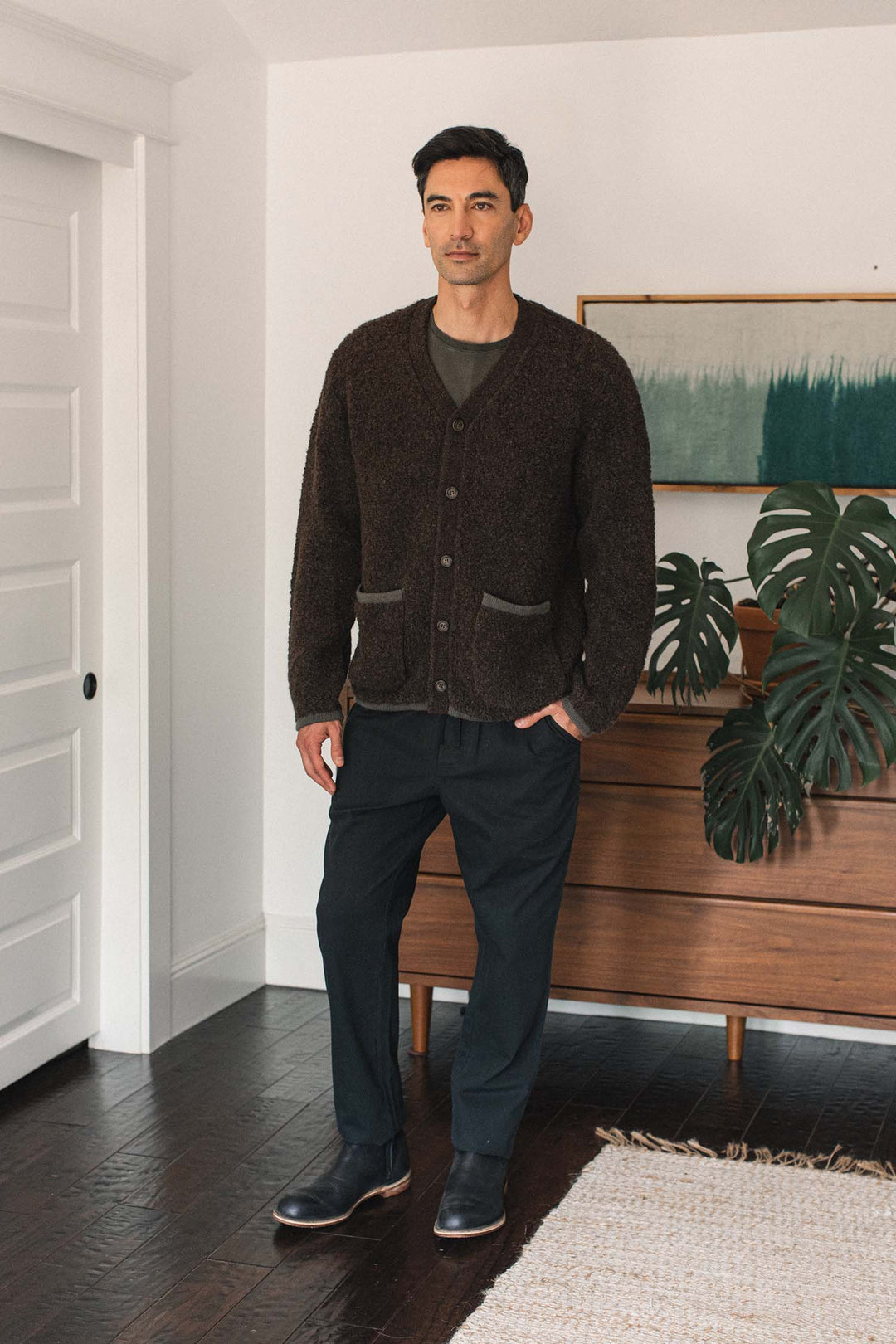 A man stands indoors on dark wood flooring, wearing the Bridge & Burn Mateo Cardigan in Dark Olive, featuring a textured knit V-neck design with patch pockets. He pairs it with dark pants and boots. Behind him are a wooden dresser, a large plant, and a framed artwork.
