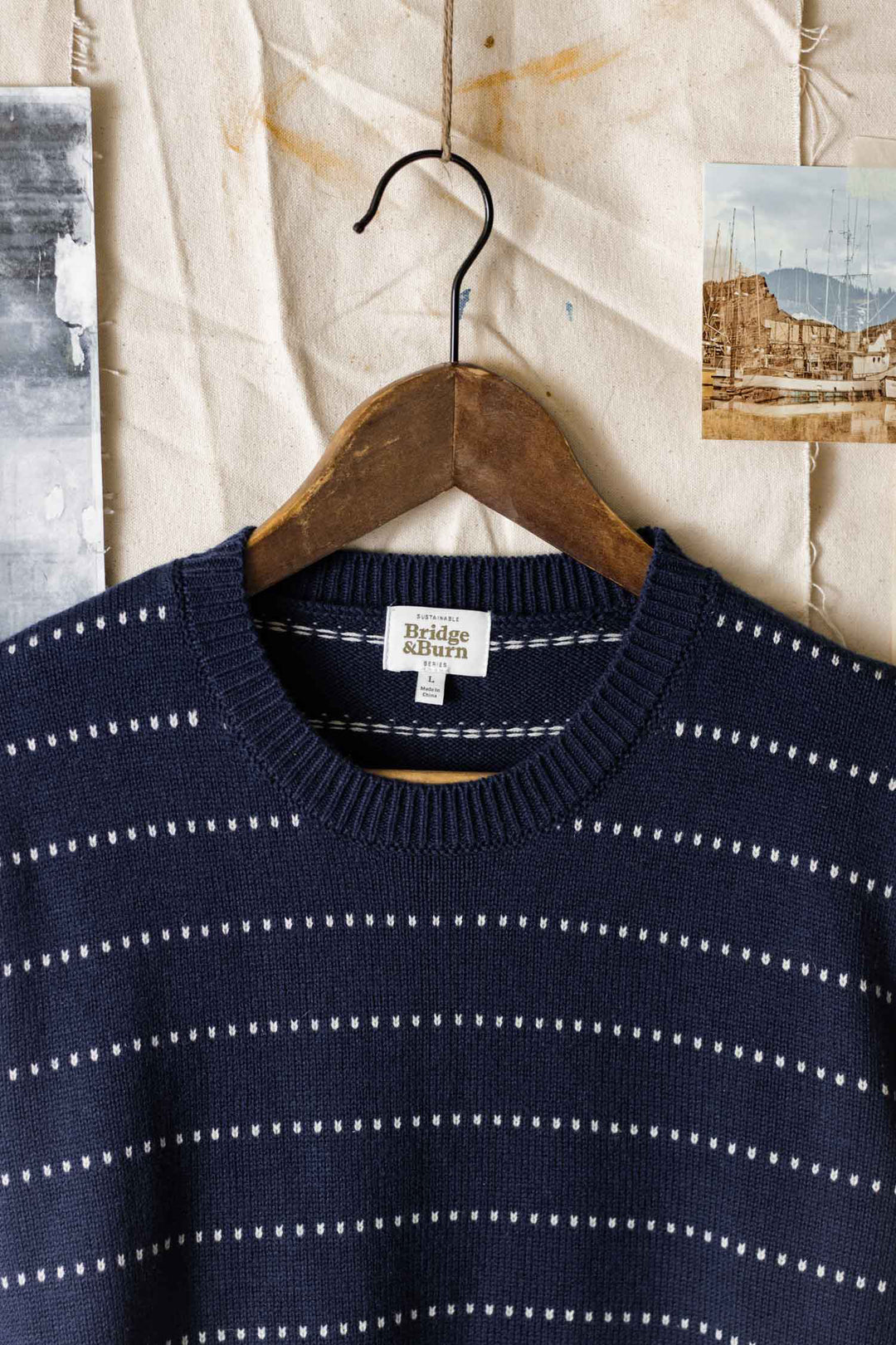 The Orson Sweater in Navy Dotted Path by Bridge & Burn hangs on a wooden hanger against a textured beige backdrop, flanked by two vintage photographs.