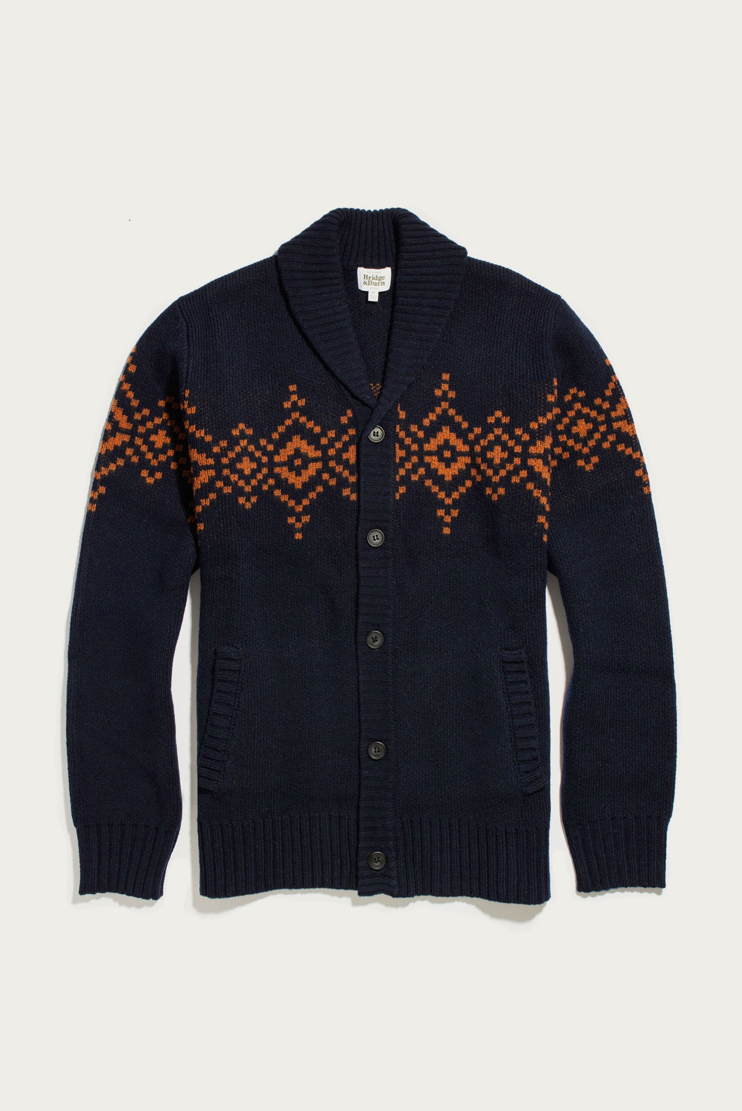 The Sprague Cardigan in Highland Dusk by Bridge & Burn is a navy blue shawl collar cardigan crafted from soft RWS wool, featuring a button closure, ribbed trim, front pockets, and an orange geometric pattern across the chest and sleeves.