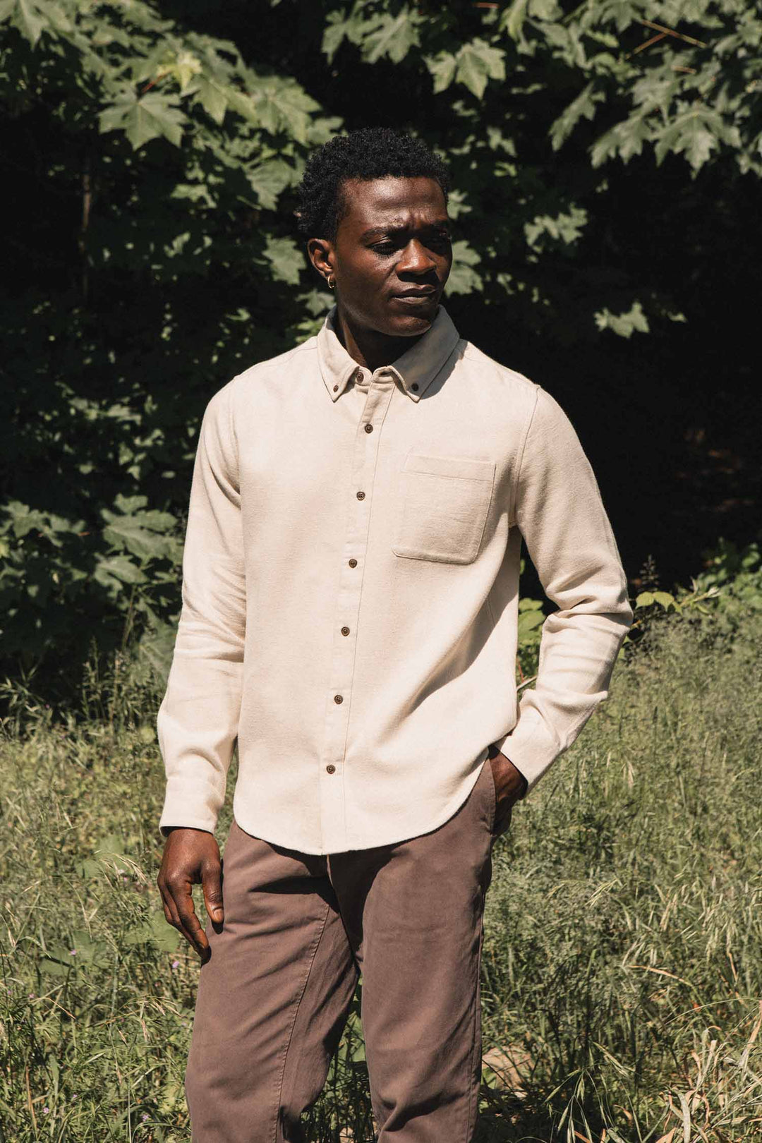 A man stands outdoors in front of green foliage, wearing the Bridge & Burn Sutton Slim Shirt in Fawn Flannel and brown pants, with one hand in his pocket, looking off to the side.