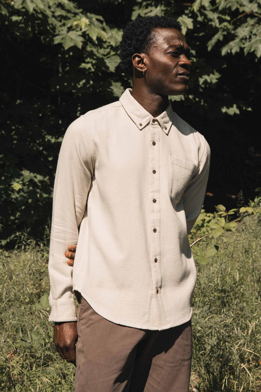 A man stands outdoors in sunlight, wearing the Bridge & Burn Sutton Slim Shirt in Fawn Flannel and brown pants, with greenery and trees in the background.