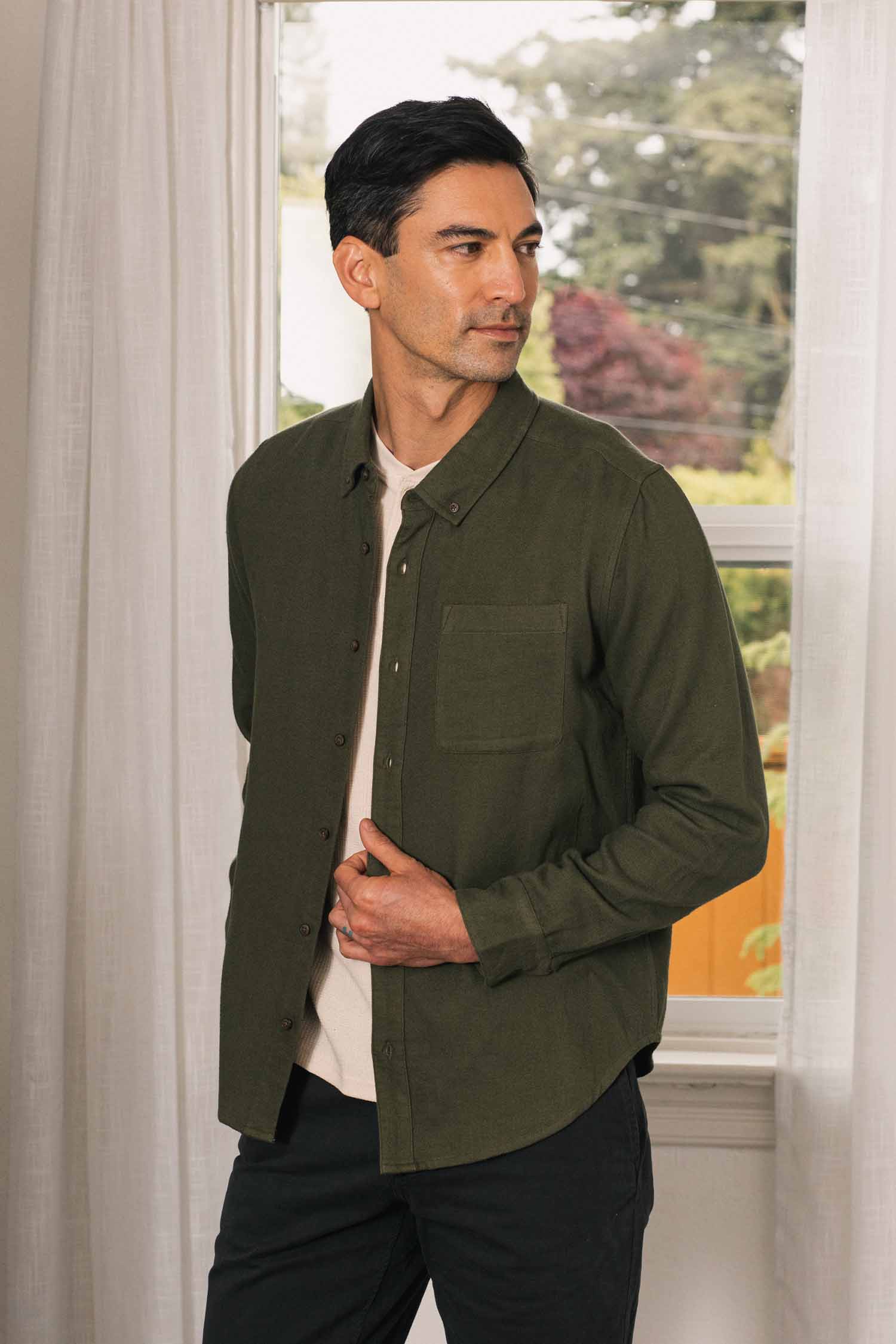 A man stands indoors by a window, wearing the Bridge & Burn Sutton Slim Shirt in Pine Flannel over a light-colored t-shirt and black pants. He looks to his left, adjusting his shirt with one hand. Trees are visible outside the window, complementing the space’s minimalist design.