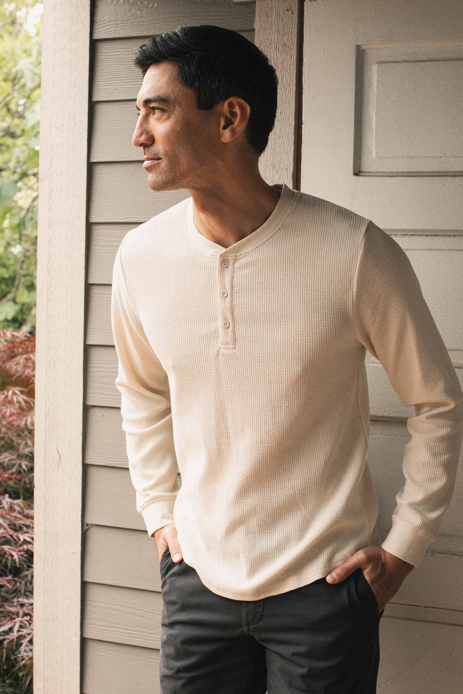 A man wearing the Bridge & Burn Men’s Recycled Waffle Henley in Ivory stands in front of a light wooden wall, looking to his left with one hand in his pocket.