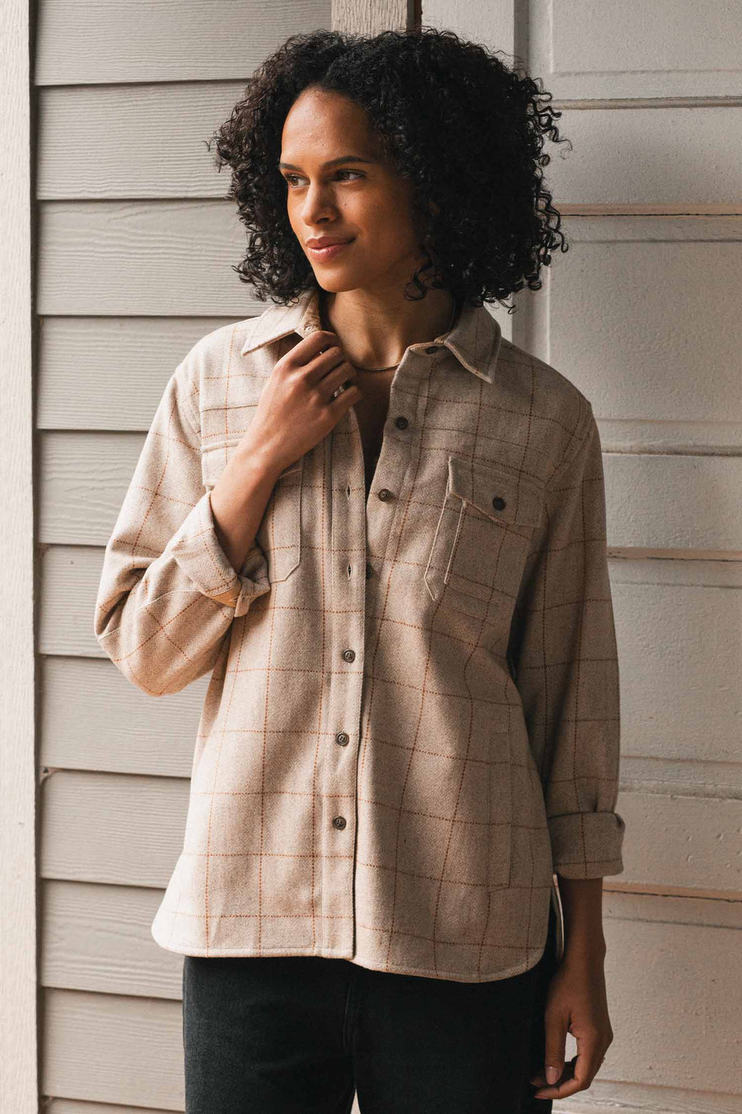 A person with curly hair stands by a light wood wall, wearing the Bridge & Burn Harlow Shirt Jacket in Stonecross and black pants, looking to the side with a gentle expression.
