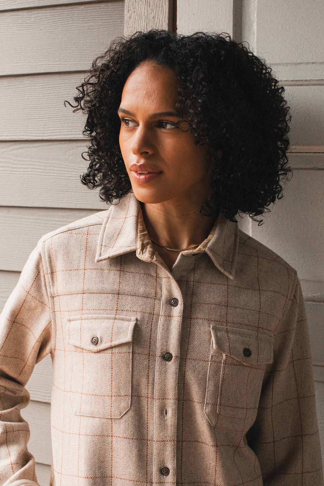 A person with curly dark hair wears the Bridge & Burn Harlow Shirt Jacket in Stonecross, standing before a light wooden wall and gazing thoughtfully to the side.