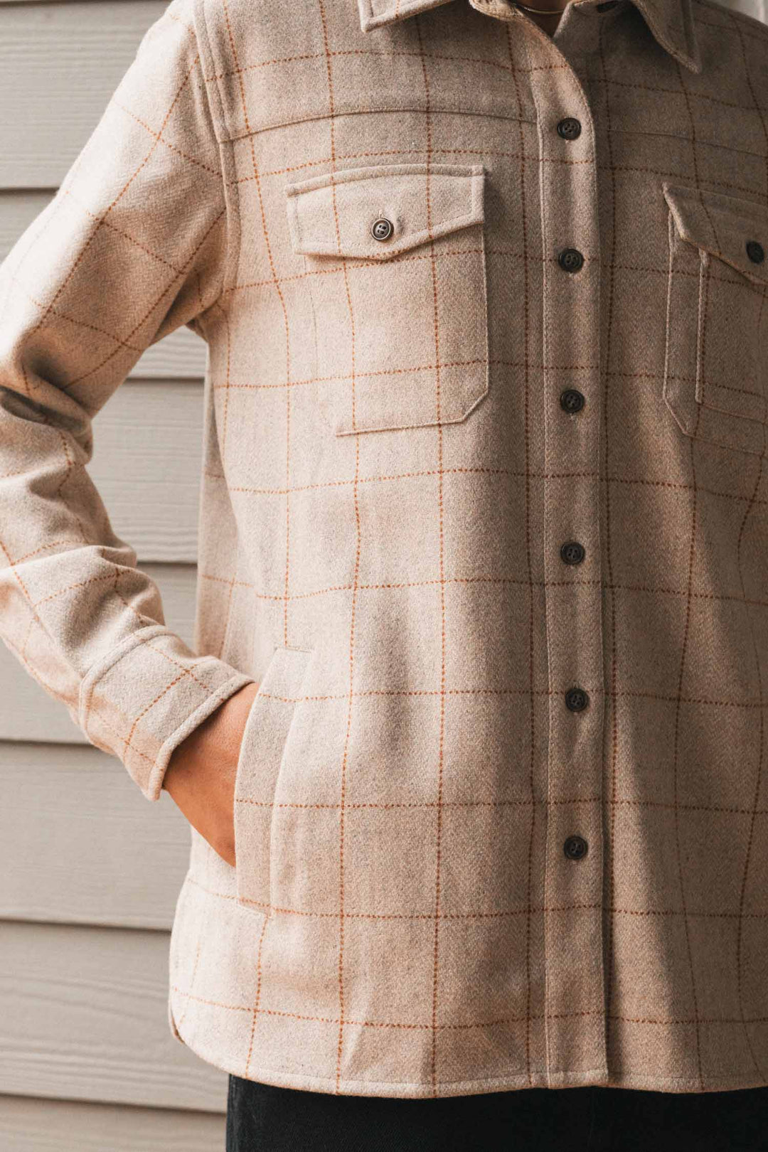 A person wearing the Bridge & Burn Harlow Shirt Jacket in Stonecross, featuring chest pockets, stands with one hand in their pocket in front of a light-colored wooden wall.