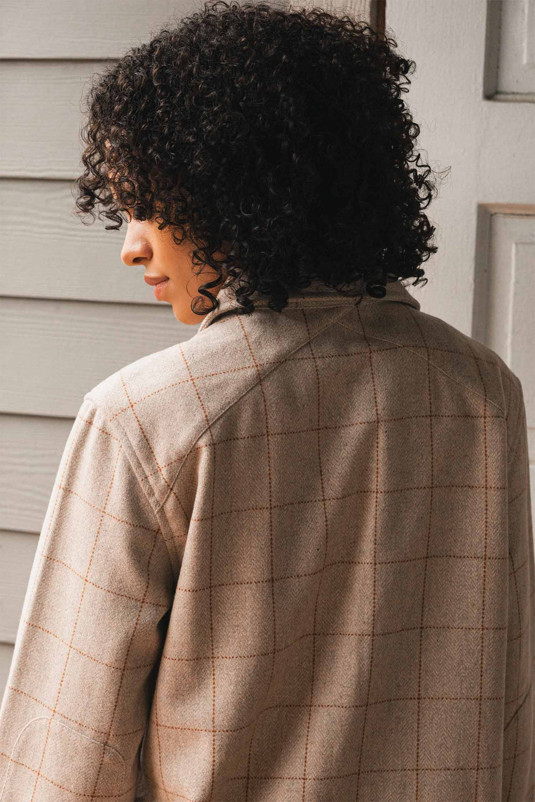 A person with curly dark hair faces away, head turned to the side, near a light wooden wall, wearing the Bridge & Burn Harlow Shirt Jacket in Stonecross.