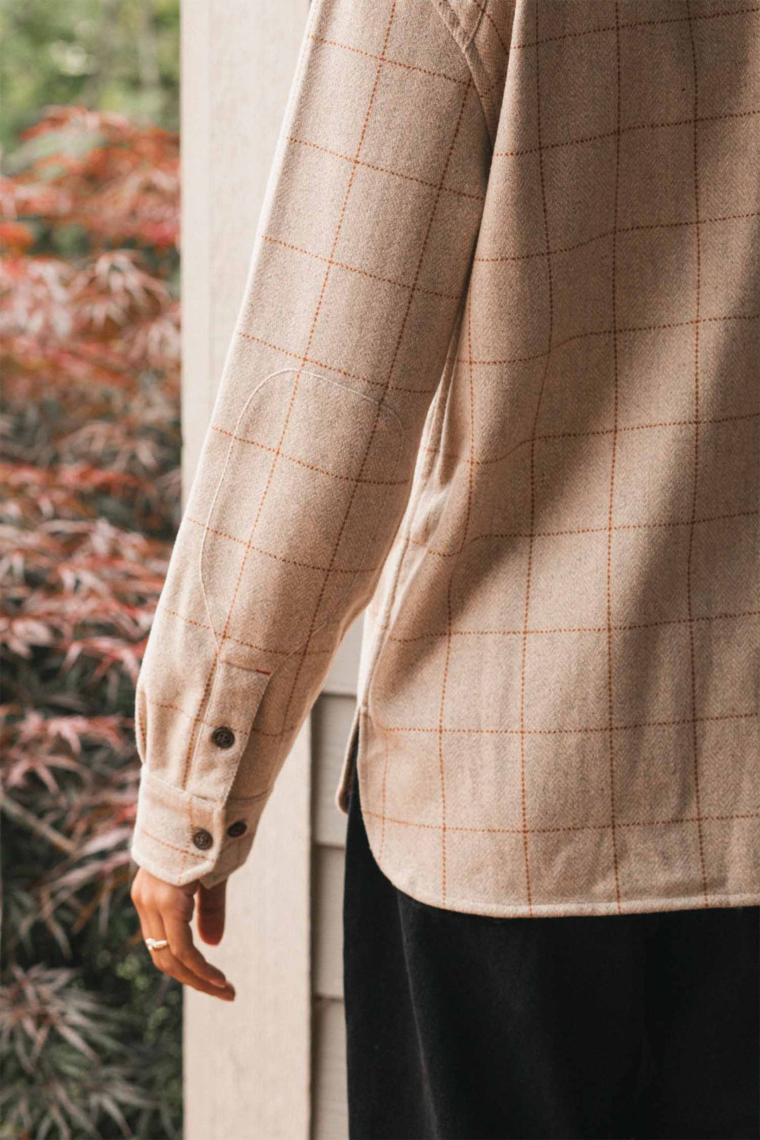 A person wearing the Bridge & Burn Harlow Shirt Jacket in Stonecross and black pants stands near a wall, showing their back and left arm. Blurred red and green plants are visible in the background.