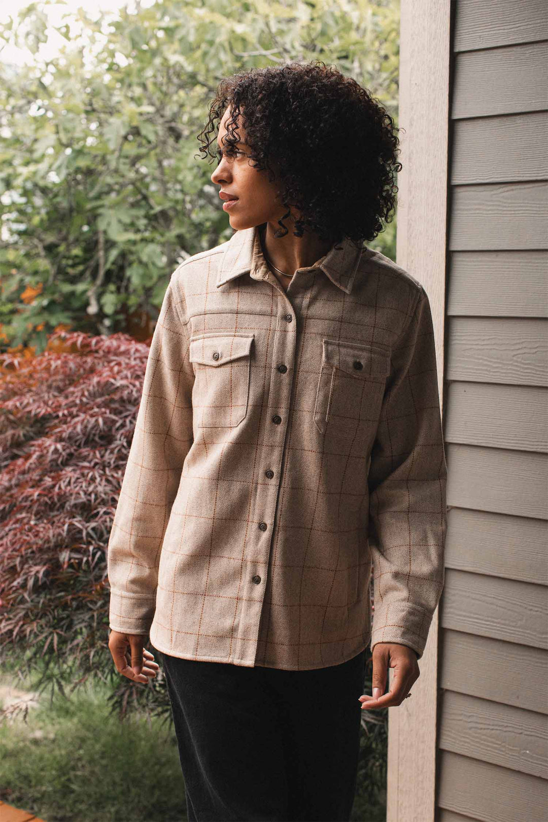 A person with curly hair stands outside near a building, wearing the Bridge & Burn Harlow Shirt Jacket in Stonecross, looking to the side. Green foliage and red plants are visible in the background.