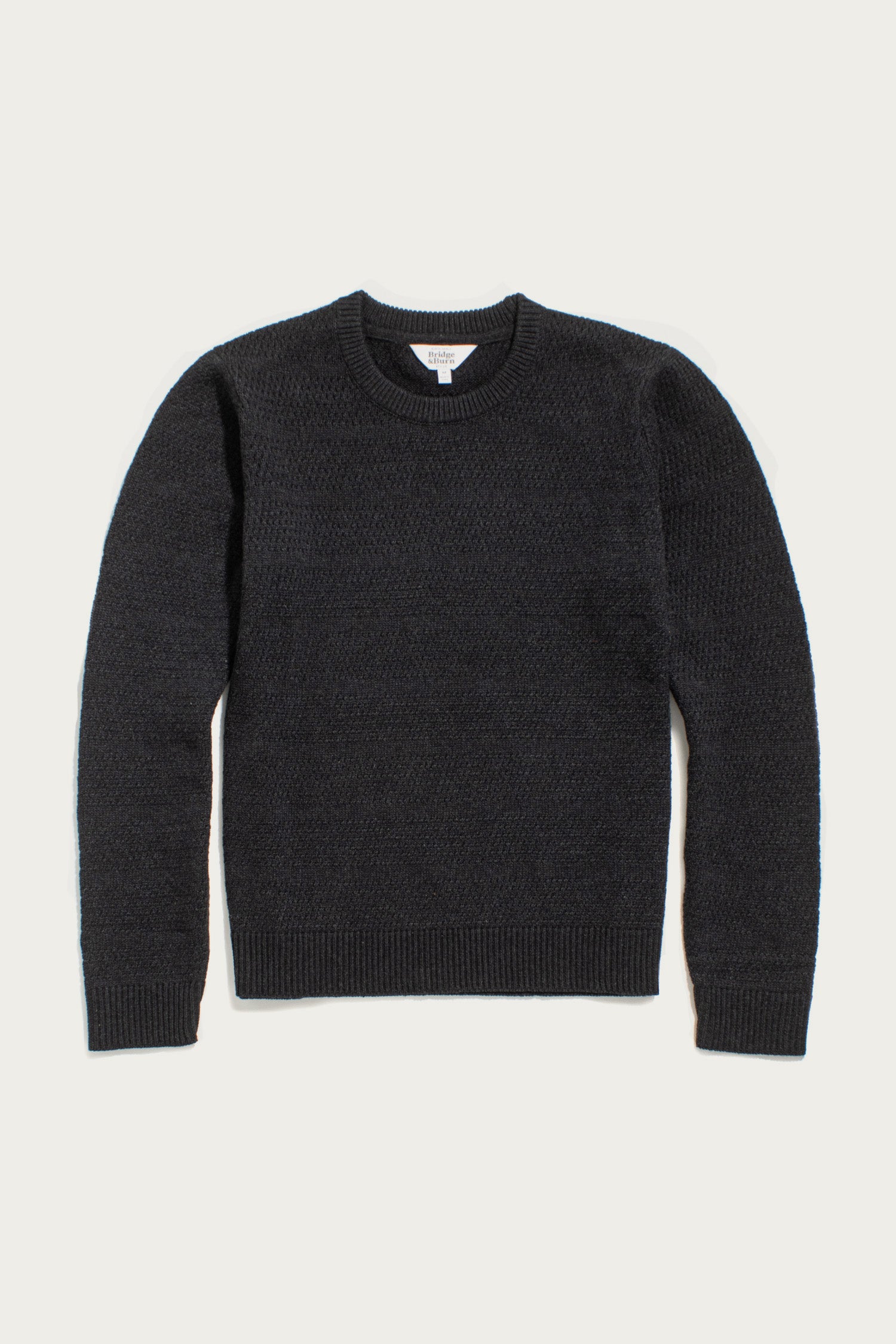 The Ansel Sweater in Midnight by Bridge & Burn is a dark gray crewneck with long sleeves, ribbed cuffs, hem, and collar, made from recycled fibers—a sustainable wardrobe essential.