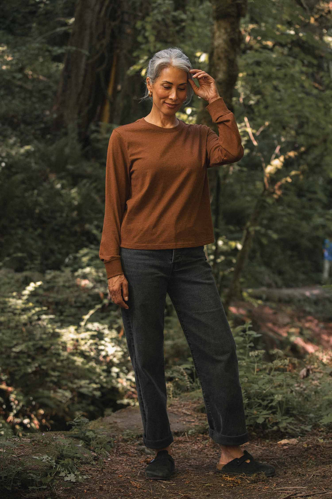 A gray-haired woman stands in a forest, wearing the relaxed fit Bridge & Burn Audrey Long Sleeve Tee in Bronze, paired with dark jeans and black shoes. With her eyes closed and a gentle smile, she softly touches her hair.