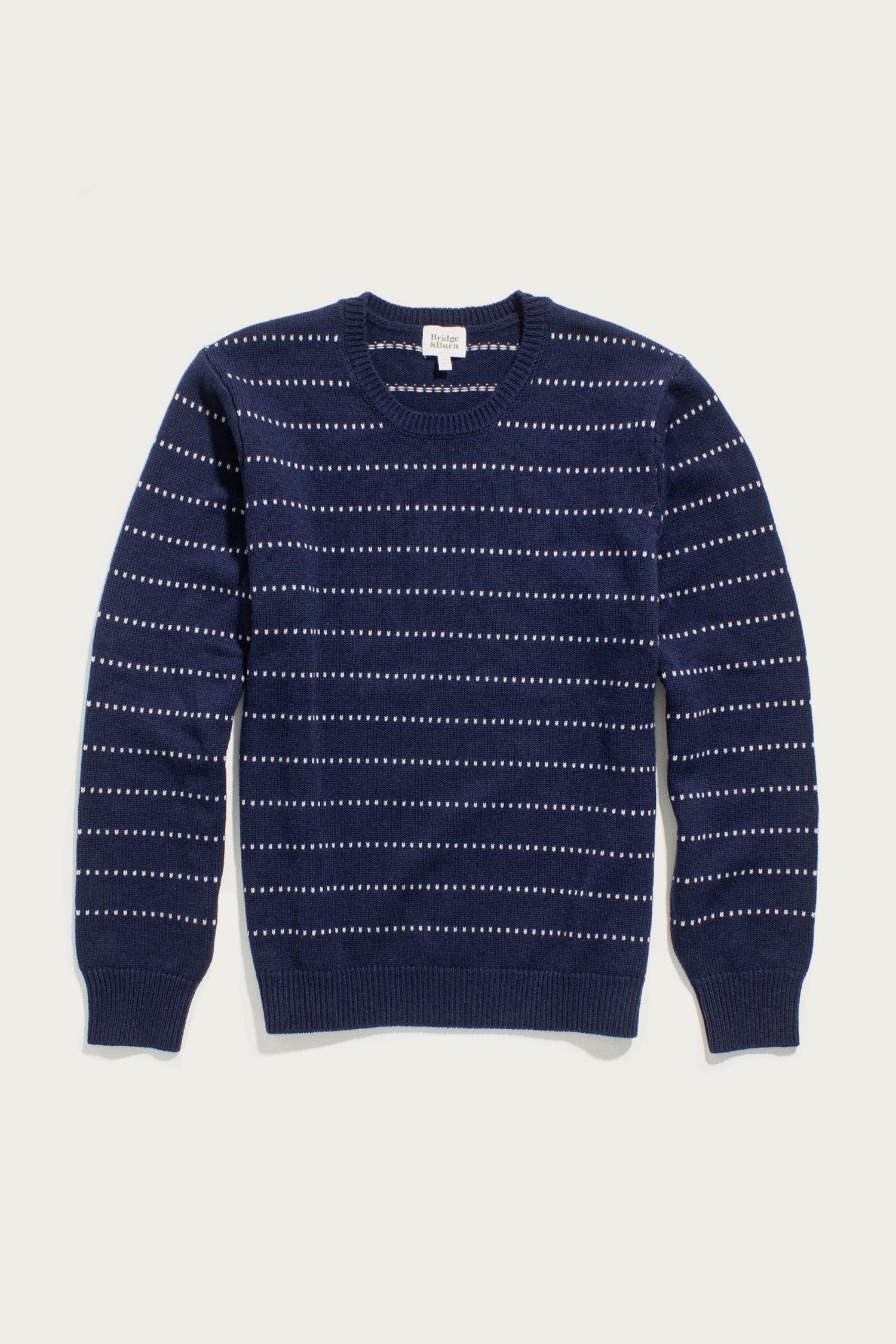 The Cline Sweater by Bridge & Burn in Navy Dotted Path features thin horizontal white dotted lines, long sleeves, and ribbed cuffs, collar, and hem, shown laid flat on a white background.