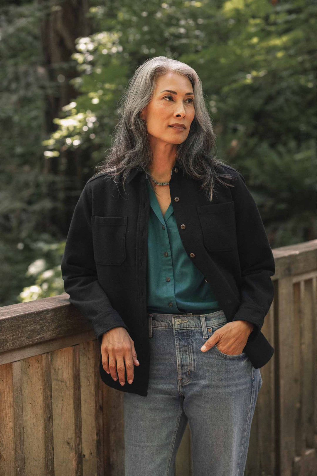 A woman with long gray hair stands outdoors by a wooden railing, looking thoughtful amid a backdrop of greenery, wearing the Bridge & Burn Dawson Overshirt in Ink Stone—crafted from recycled cotton—layered over a teal shirt and jeans.