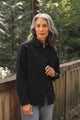 A woman with long gray hair, dressed in the Bridge & Burn Dawson Overshirt in Ink Stone and jeans, stands outdoors on a wooden bridge with a forest backdrop.