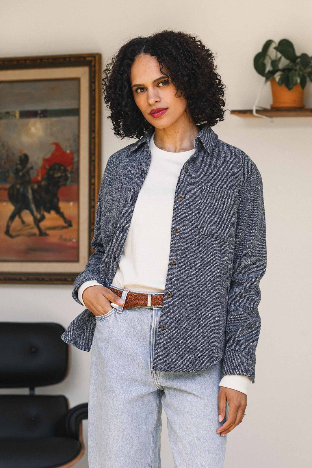 A woman with curly hair wearing Bridge & Burn’s Dawson Overshirt in Ocean Marl and light jeans stands indoors near a painting and a shelf with a potted plant, embodying a sustainable choice in fashion.