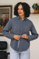 A person with curly hair wearing the Bridge & Burn Dawson Overshirt in Ocean Marl and light jeans stands indoors, tying the waist of their shirt. A painting and a potted plant in the background highlight their sustainable choice.