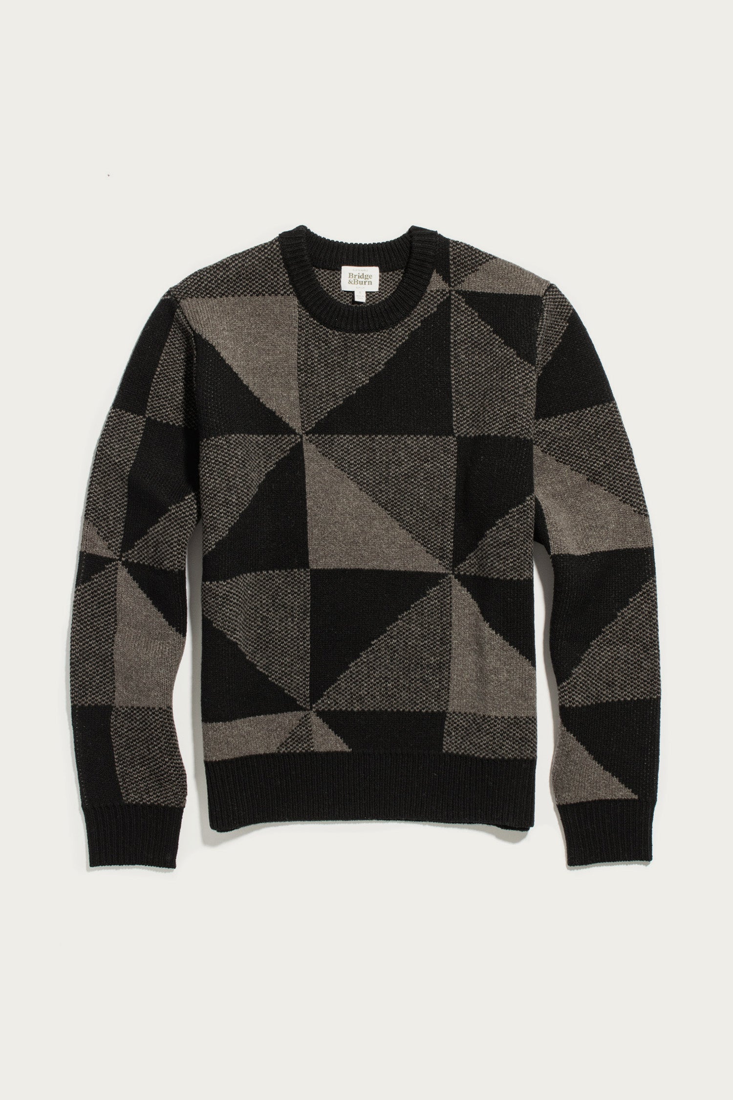 The Bridge & Burn Hollis Sweater / Stone Gale is a geometric-patterned crew neck sweater in black and gray, featuring triangles and rectangles. Crafted from soft Merino wool for style and comfort, it includes long sleeves with ribbed cuffs, collar, and hem, shown laid flat on a white background.