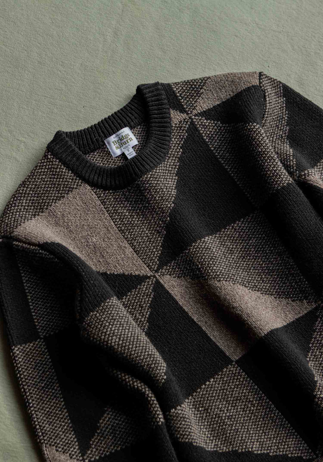 A Bridge & Burn Hollis Sweater in the Stone Gale colorway, featuring a black and brown geometric pattern in Merino Wool, is laid flat on a green surface to display a close-up of the front and crew neck collar with the Hollis Sweater label visible.