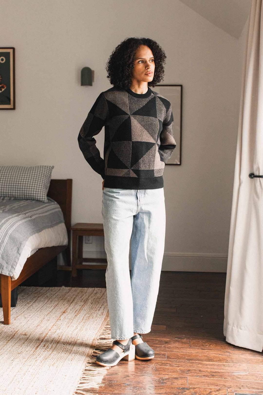 A person with curly hair stands on a rug in a bedroom, wearing the Bridge & Burn Hollis Sweater in Stone Gale—a geometric-patterned crew neck sweater made from soft merino wool—paired with light-wash jeans and clogs. A made bed and artwork are visible in the background.