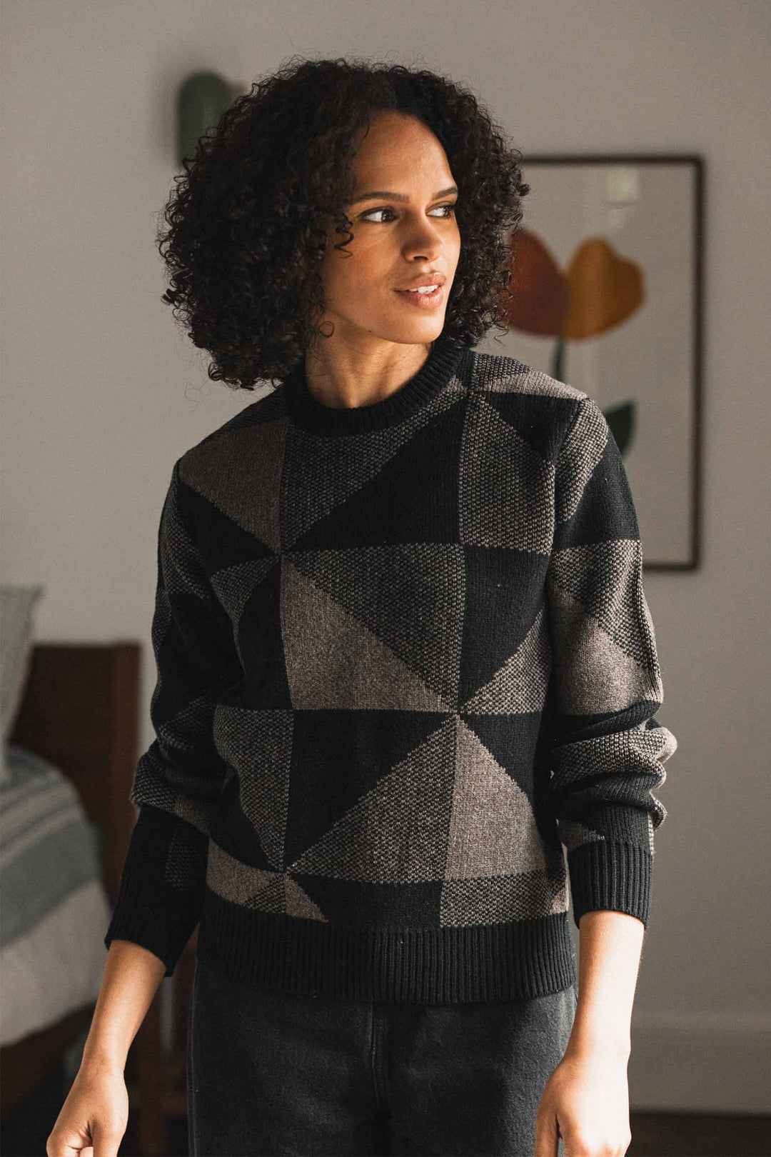 A person with curly hair wears the Bridge & Burn Hollis Sweater in Stone Gale, standing indoors beside a bed with framed abstract art on the wall in the background.