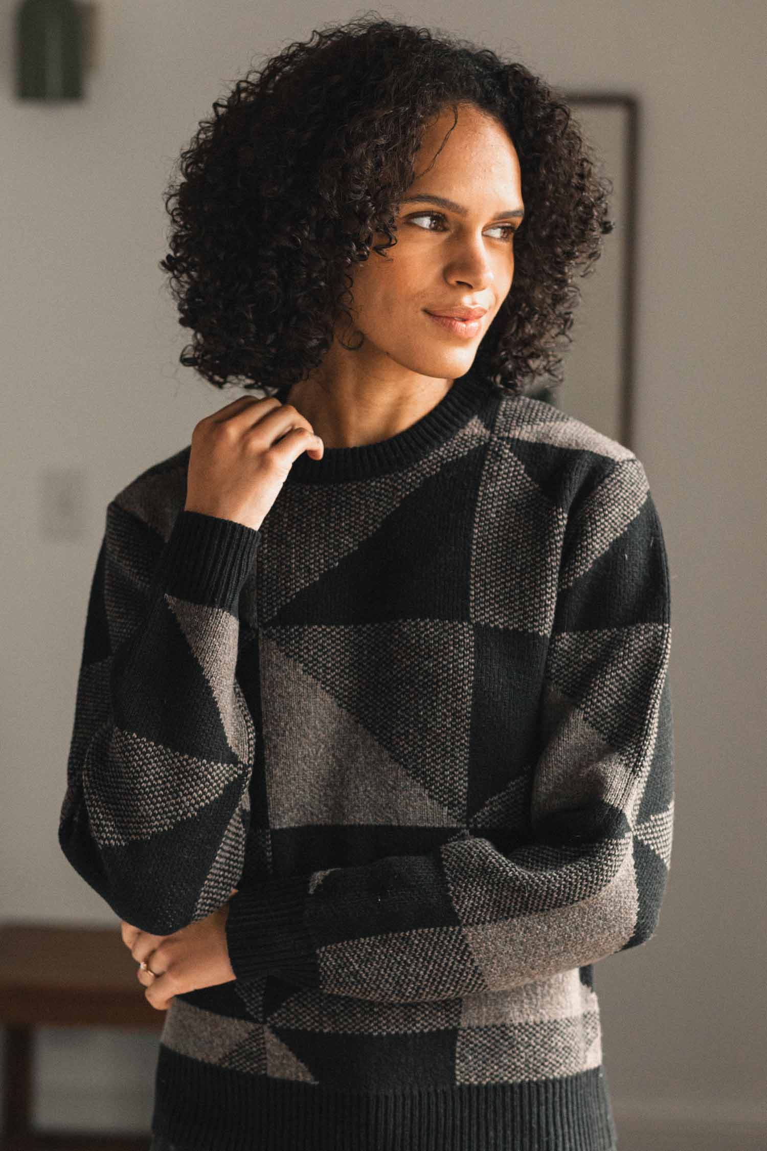 A woman with curly hair stands indoors, gazing thoughtfully to the side while wearing the Bridge & Burn Hollis Sweater in Stone Gale—a geometric-patterned black and gray crew neck.