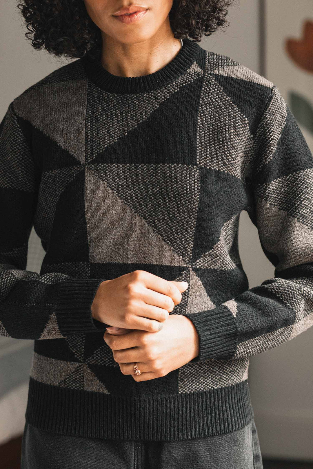 A person wearing the Bridge & Burn Hollis Sweater in Stone Gale, featuring a geometric pattern in black and gray, stands indoors with their hands clasped together. Their face is partially visible, and they have curly dark hair.