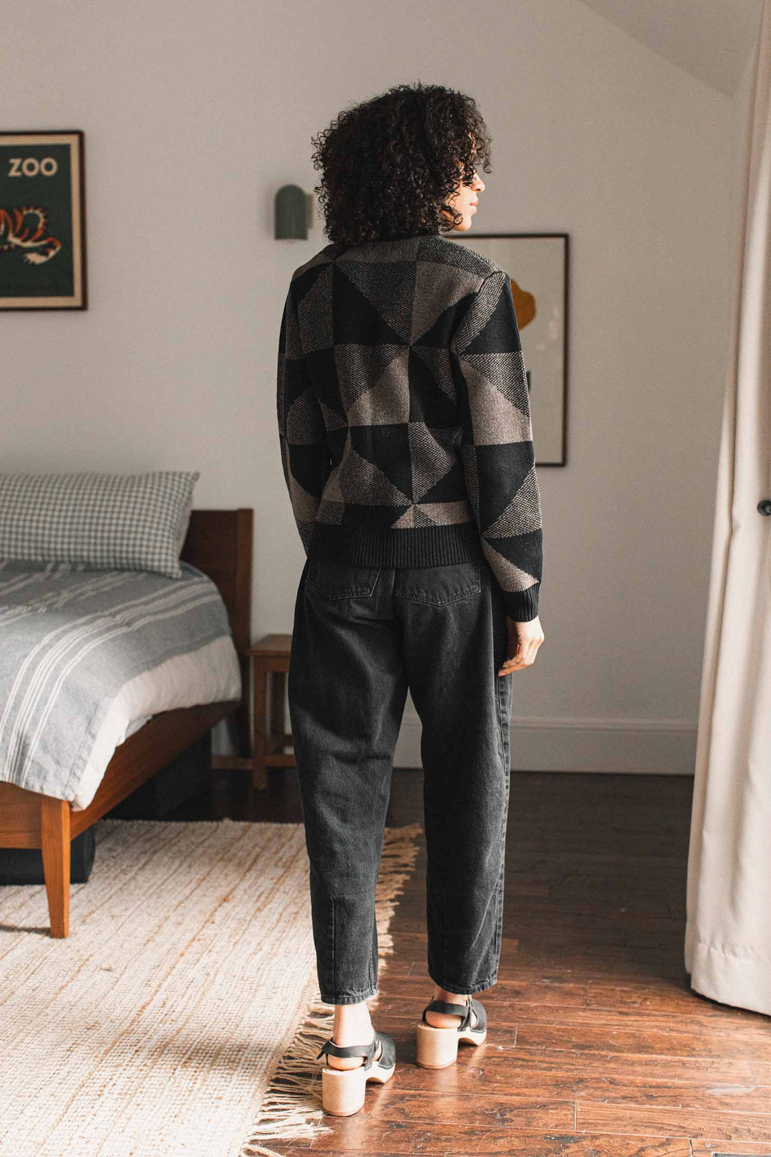 A person with curly hair stands in a bedroom, facing away. They wear the Bridge & Burn Hollis Sweater in Stone Gale, paired with black jeans and sandals. The room features a bed, wooden furniture, and framed art on white walls.
