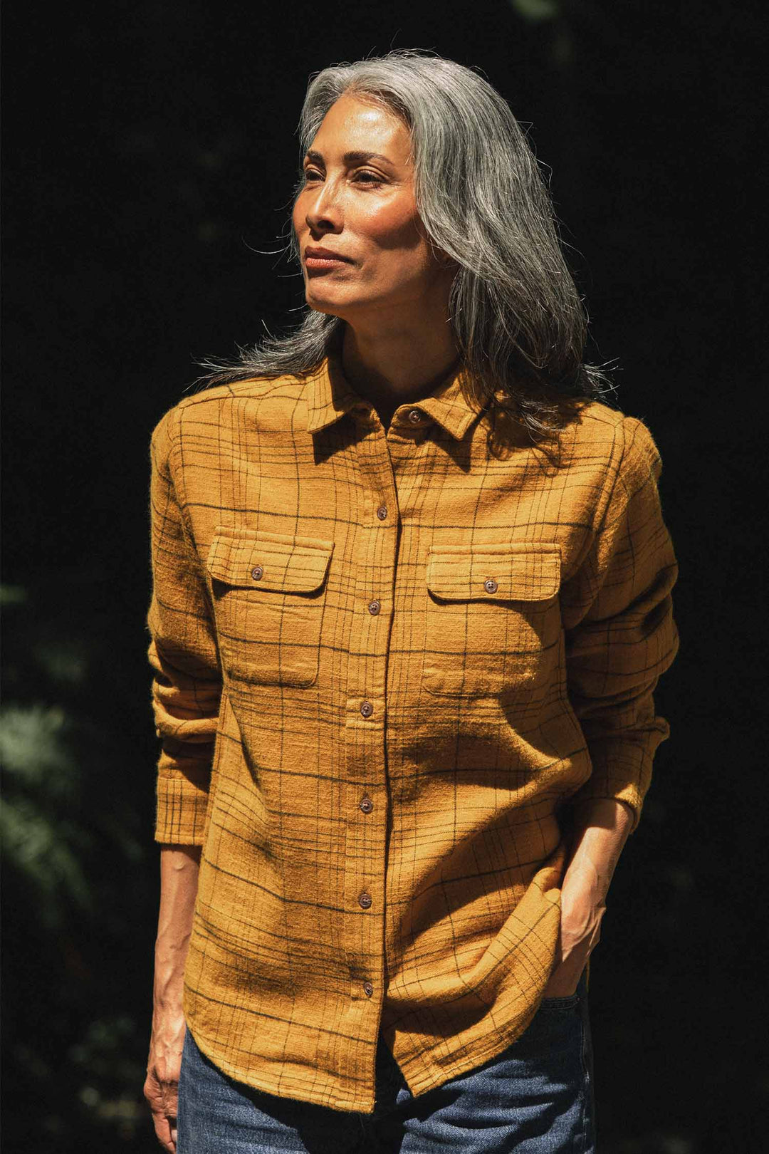 A woman with long gray hair stands outdoors in sunlight, wearing the Bridge & Burn Lenon Shirt in Golden Horizon Plaid and jeans, with one hand in her pocket against a dark background.