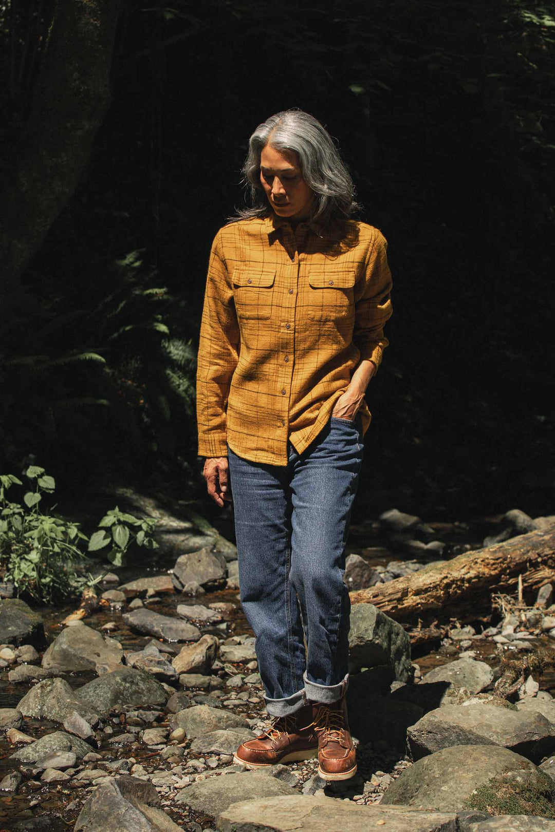 A person with gray hair, wearing the Bridge & Burn Lenon Shirt in Golden Horizon Plaid, blue jeans, and brown boots, walks on rocks amid sunlit forest plants and fallen branches.