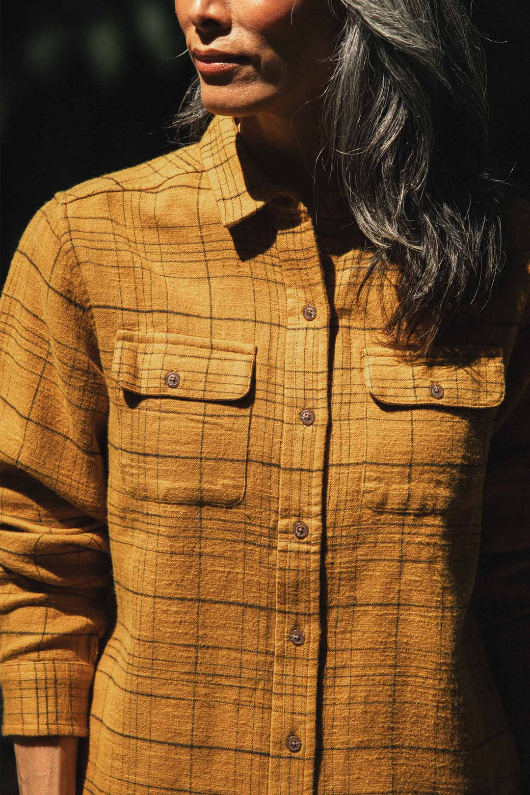 A person with long gray hair stands in sunlight, wearing the Bridge & Burn Lenon Shirt in Golden Horizon Plaid. The cropped image highlights its organic cotton texture, standard fit, and practical pockets.