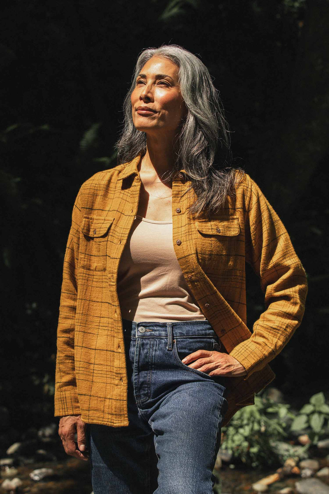 A woman with long gray hair stands outdoors in sunlight, wearing the Bridge & Burn Lenon Shirt in Golden Horizon Plaid over a beige top and blue jeans, with one hand on her hip and a thoughtful expression.