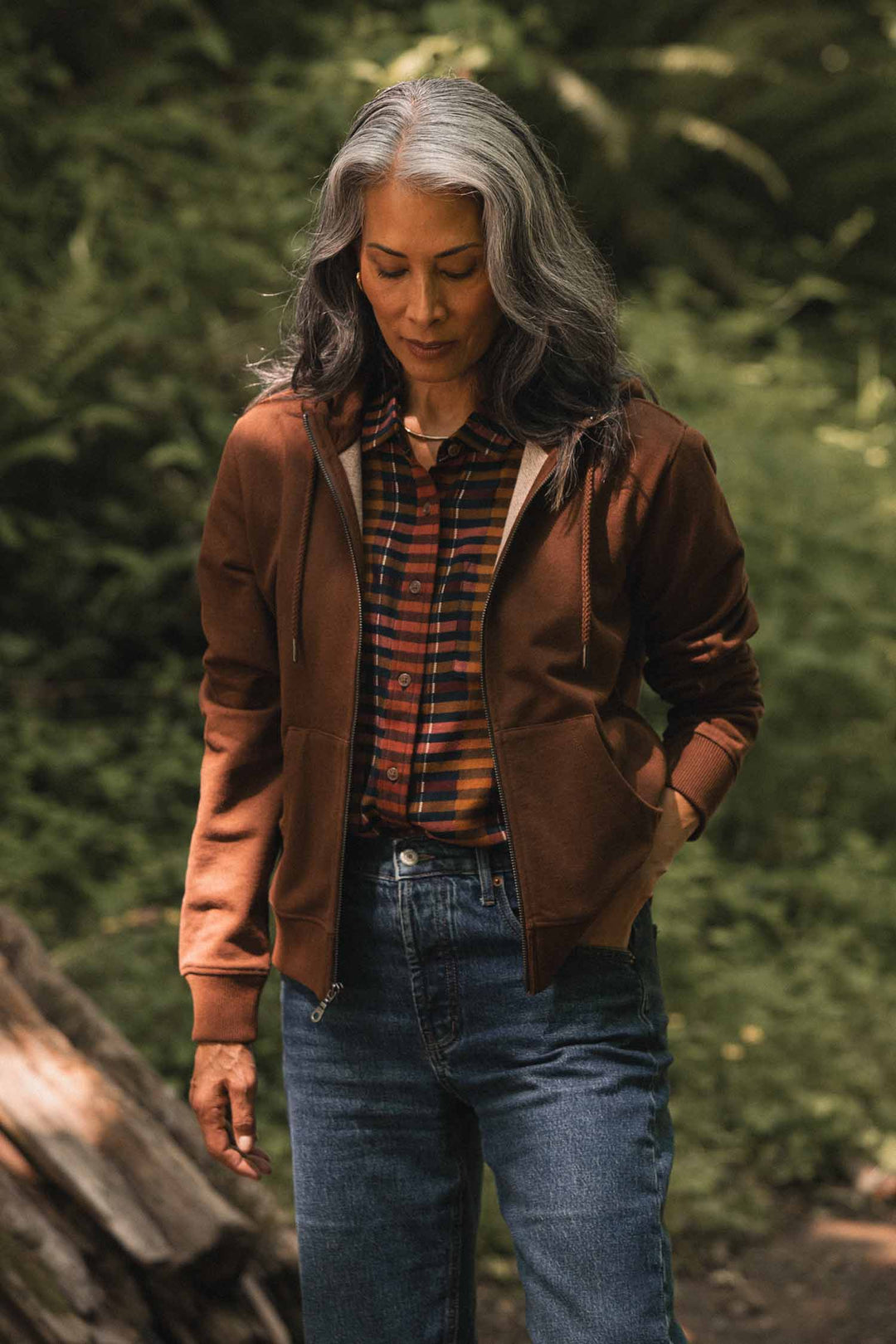 A woman with long gray hair stands outdoors, surrounded by greenery, wearing the Bridge & Burn Mason Hoodie in Cumin—a brown zip-front hooded sweatshirt layered over a plaid shirt and blue jeans, her hands tucked in her pockets.