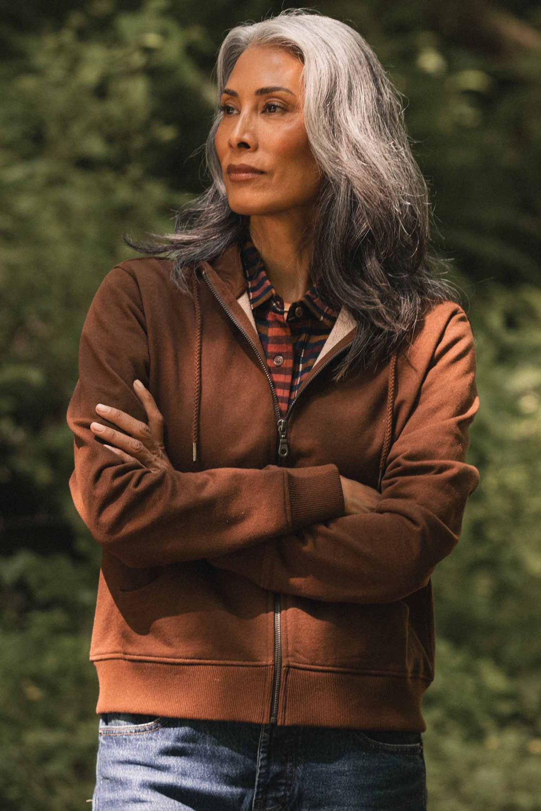 A woman with long gray hair stands outdoors in the Bridge & Burn Mason Hoodie in Cumin, a brown recycled cotton zip-up jacket, layered over a plaid shirt. With her arms crossed and a thoughtful expression as she looks to the side, blurred green foliage forms the background.