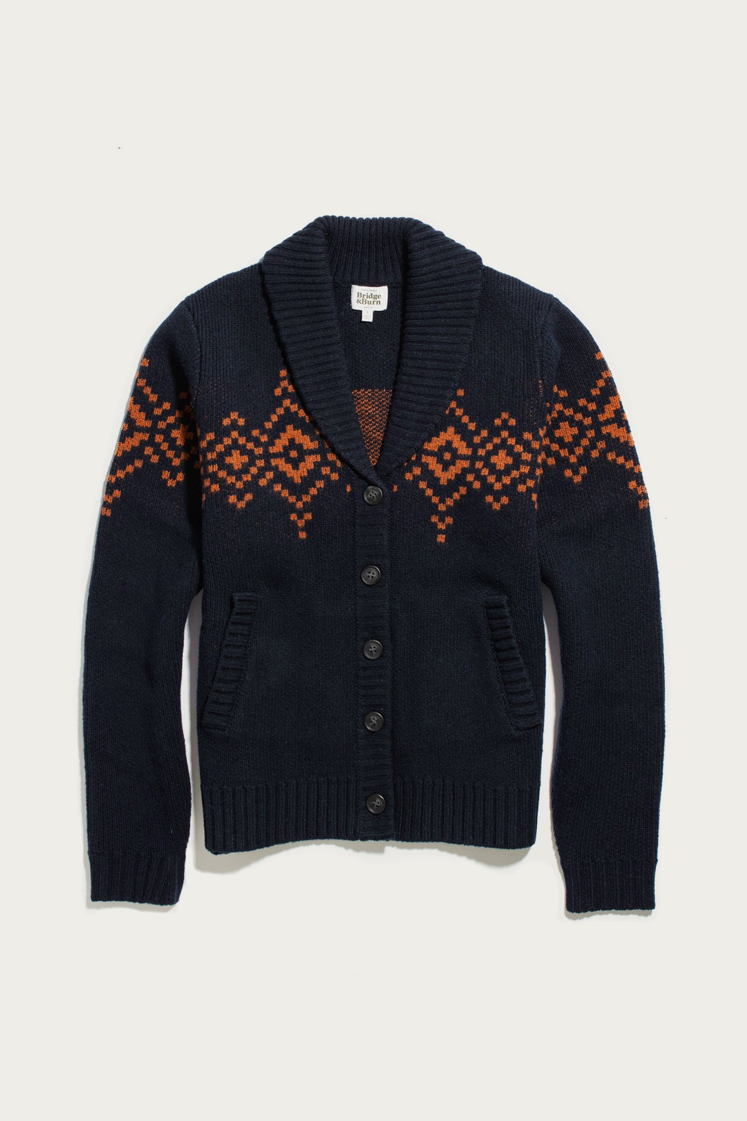 The Bridge & Burn Nolan Cardigan in Highland Dusk features a classic shawl collar, front buttons, and two pockets, with a striking orange geometric pattern across the chest and sleeves. Crafted from cozy wool in navy blue, this cardigan seamlessly combines timeless style with modern comfort.