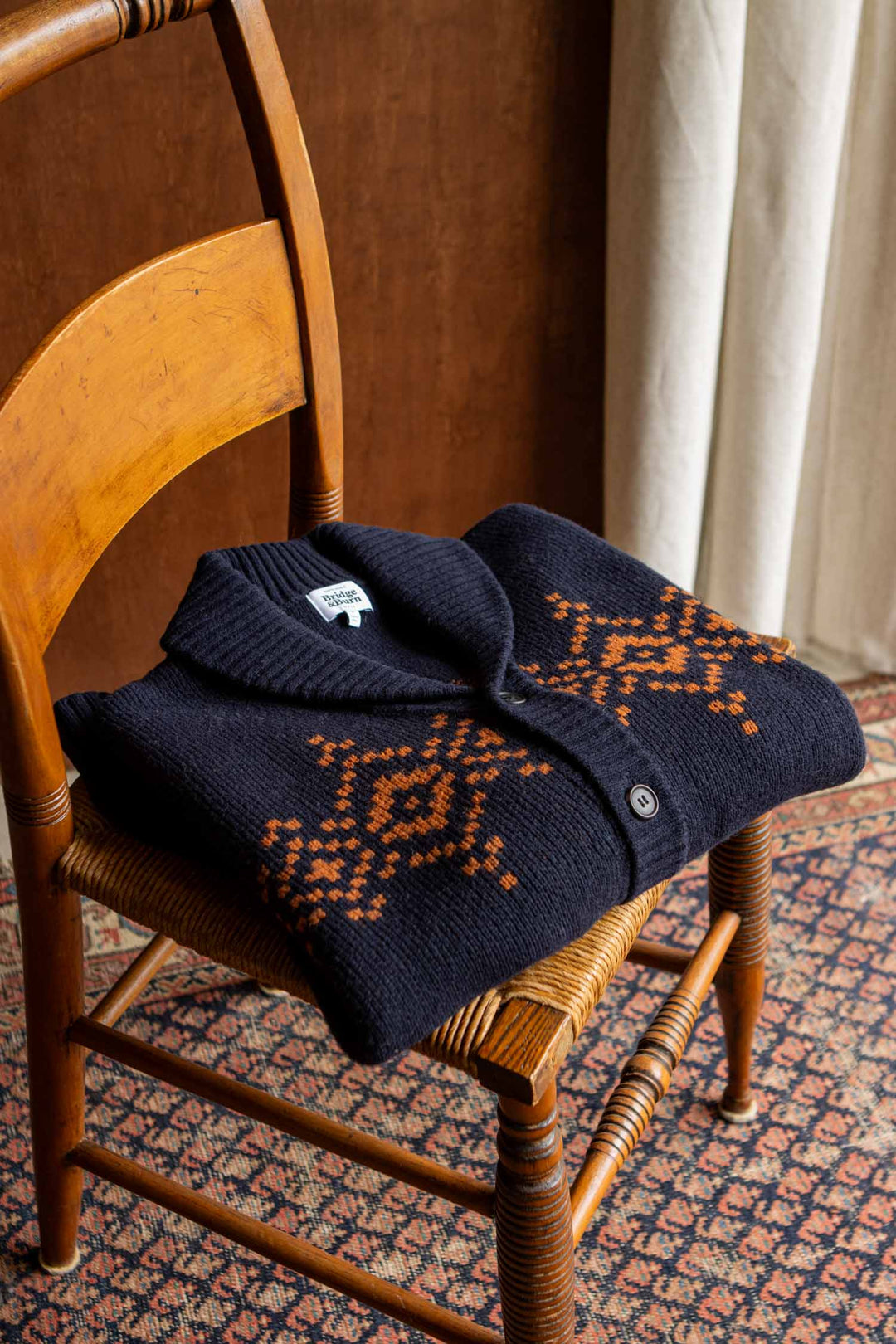The Nolan Cardigan in the Highland Dusk colorway by Bridge & Burn, featuring orange geometric patterns on navy blue wool, is folded and placed on a wooden chair with a woven seat. In the background, a white curtain hangs beside a brown wall, while a patterned rug lies beneath the chair.