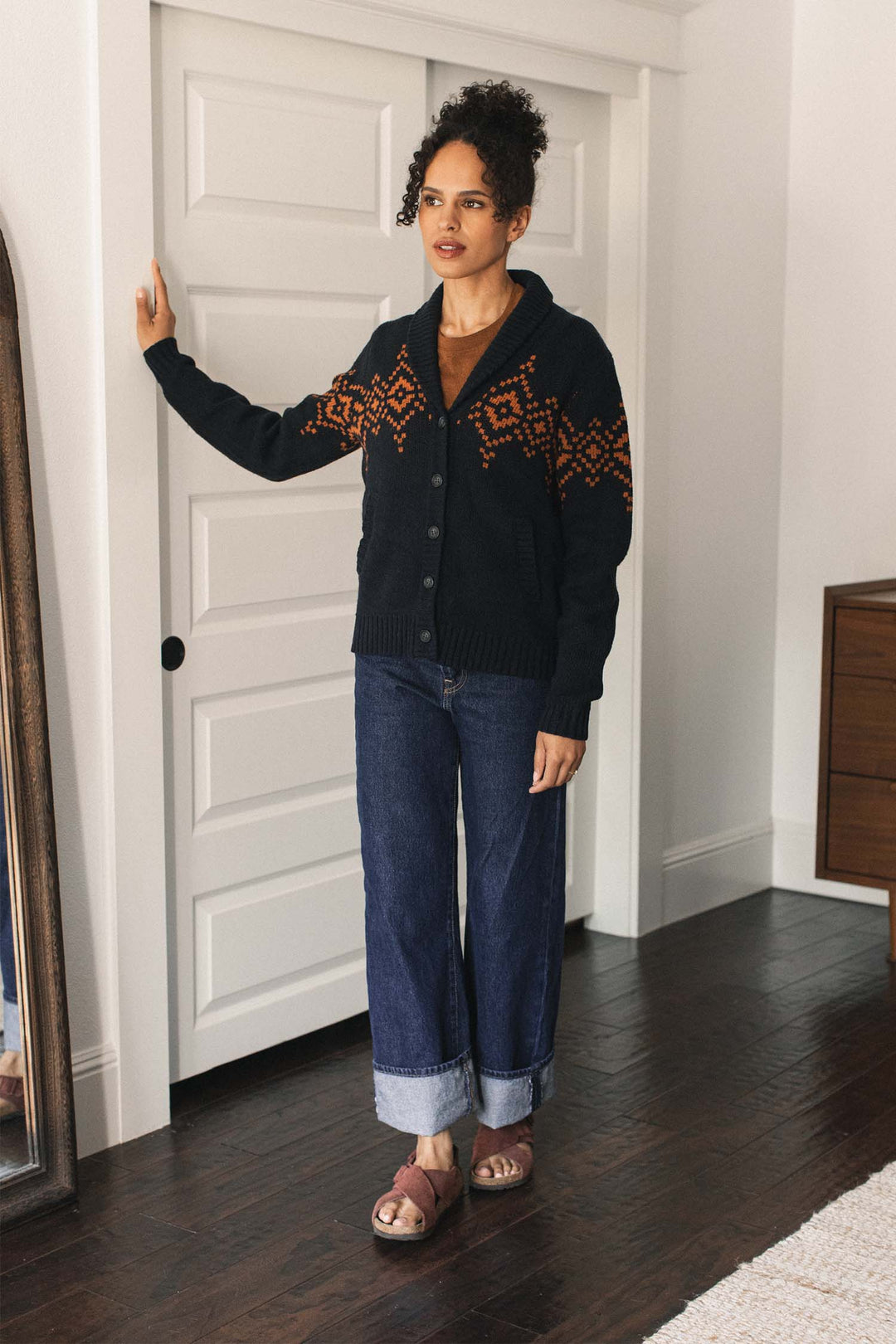 A person stands indoors by a white sliding door, wearing the Bridge & Burn Nolan Cardigan in Highland Dusk with orange patterns, cuffed jeans, and brown sandals, with one hand resting on the door frame.