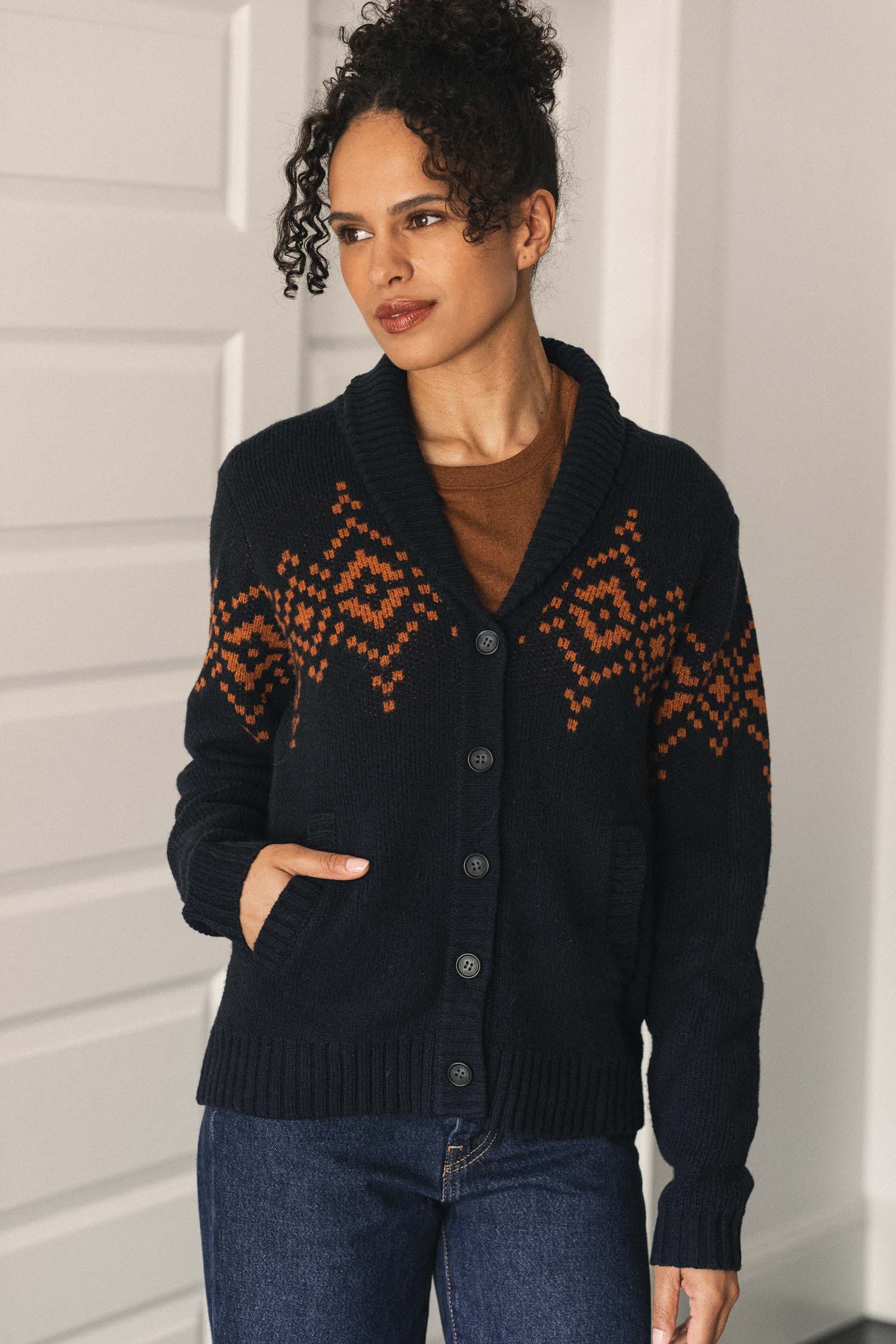A woman with curly hair stands indoors with one hand in her pocket, wearing blue jeans and the Nolan Cardigan in Highland Dusk by Bridge & Burn—a dark wool cardigan featuring an orange geometric pattern.