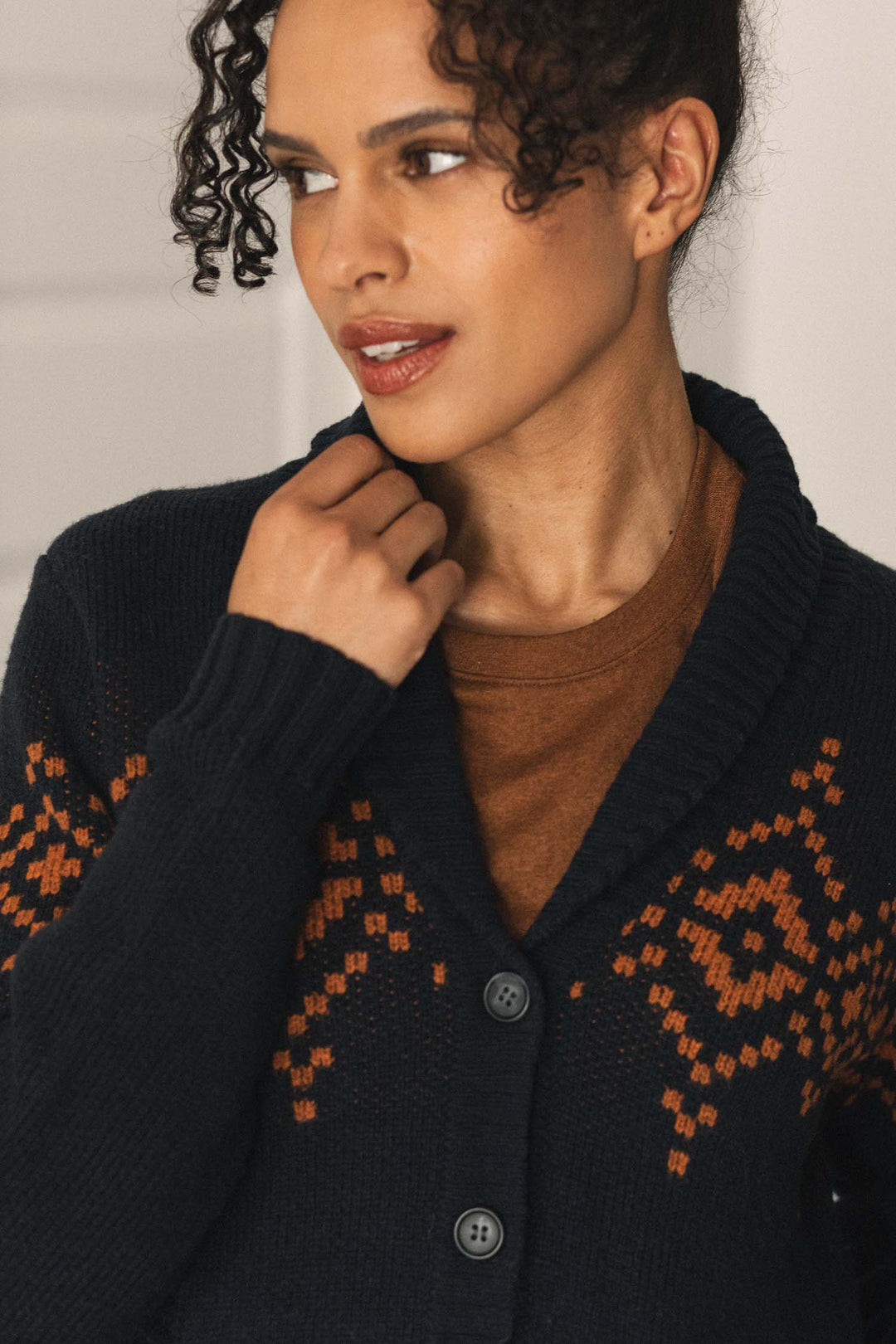 A woman with curly hair wears the Bridge & Burn Nolan Cardigan in Highland Dusk—a navy wool cardigan featuring orange patterns and a cozy shawl collar—layered over a brown shirt, as she looks to the side and gently touches her collar.