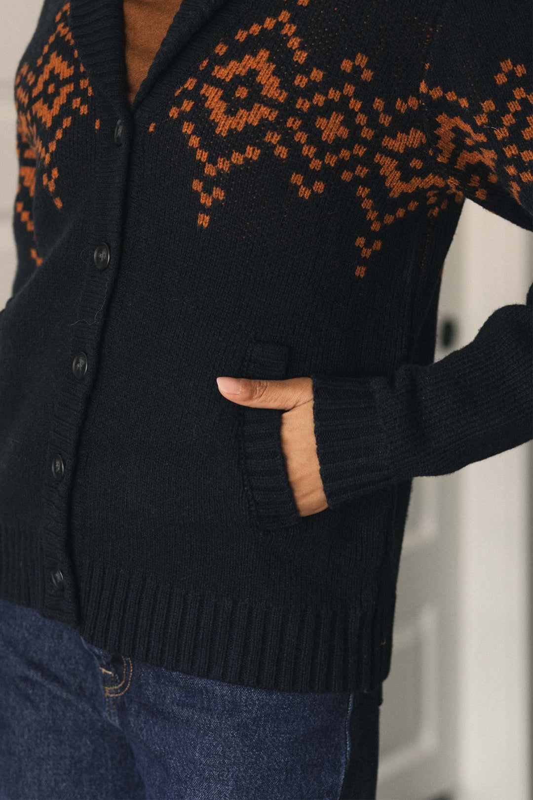 A person wearing the Bridge & Burn Nolan Cardigan in Highland Dusk, which features orange patterns and buttons, stands indoors with one hand in their pocket. The wool cardigan is paired with blue jeans.