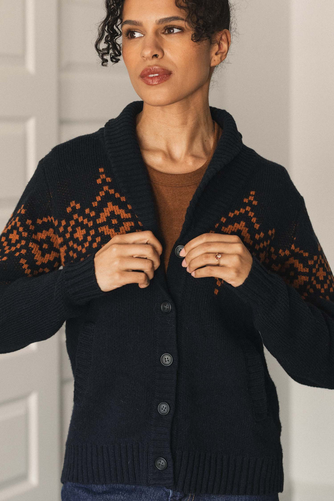 A woman with curly hair wears the Bridge & Burn Nolan Cardigan in Highland Dusk—a dark shawl collar cardigan featuring an orange geometric pattern on the sleeves—layered over a brown shirt, standing indoors with white paneled doors in the background.